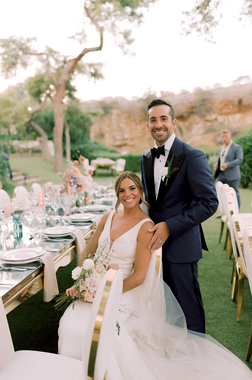A smiling bride in a white V-neck gown sits at a long outdoor wedding table on a lawn, holding a bouquet of white and blush roses, peonies and wispy greenery, while her groom in a navy suit and black bow tie stands behind her with his hand on her shoulder. The table is set with white plates, gold-rimmed chairs and tall pink taper candles among soft peach and lilac floral arrangements. These are wedding flowers in Greece, at Lake Vouliagmeni, with a rocky hillside and trees softly blurred behind them.