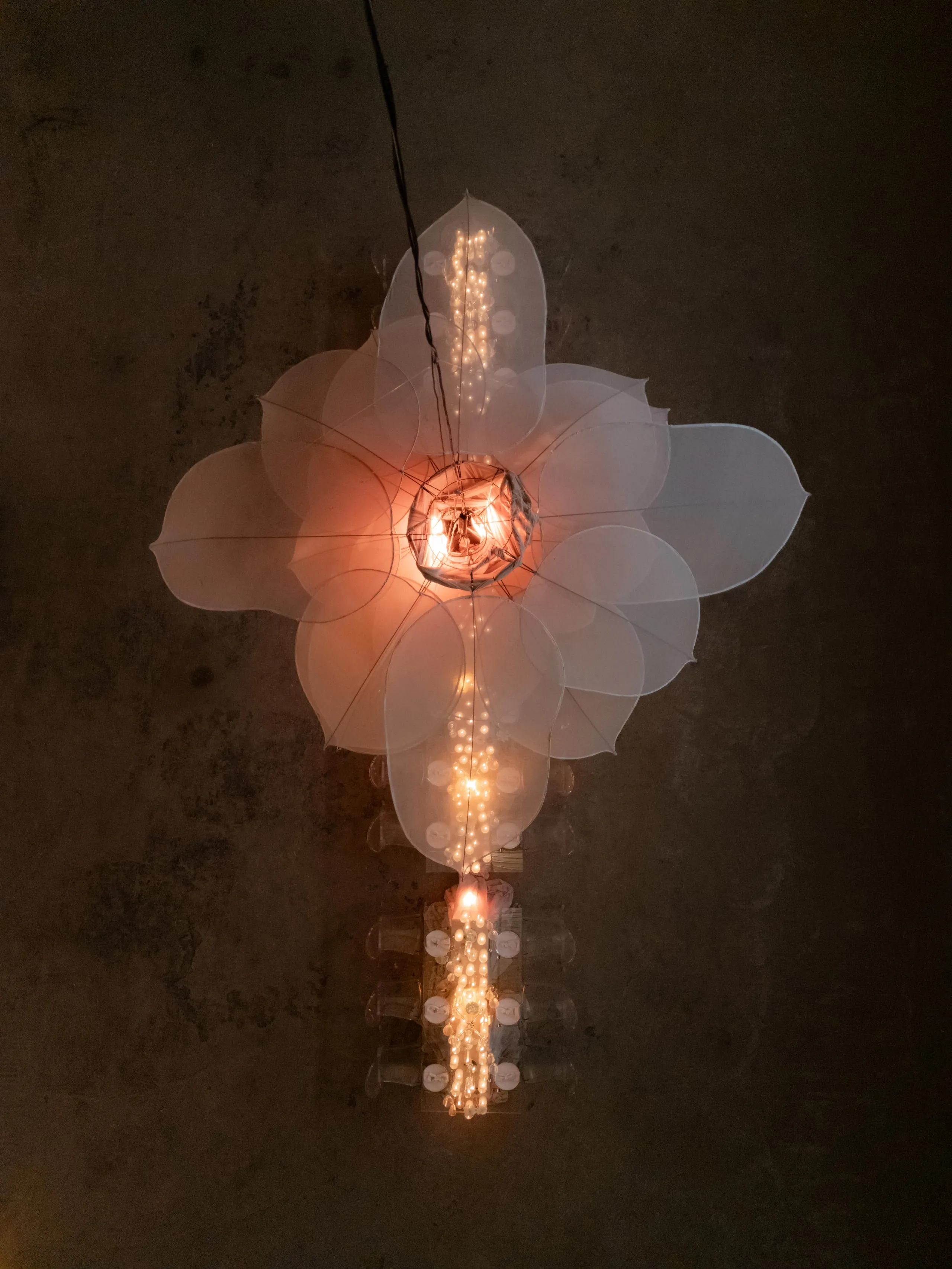 A glowing, sculptural flower hangs against a dark, textured wall, its translucent pale petals layered in overlapping rings around a warm, bulb-lit centre. Strings of tiny lights trail downward from the petals and a cluster of clear beads and plastic pieces dangles below, with a faint glow spilling through the whole form. This is a decorative light installation rather than a real floral arrangement, shown here in Rethimno, Crete, Greece.