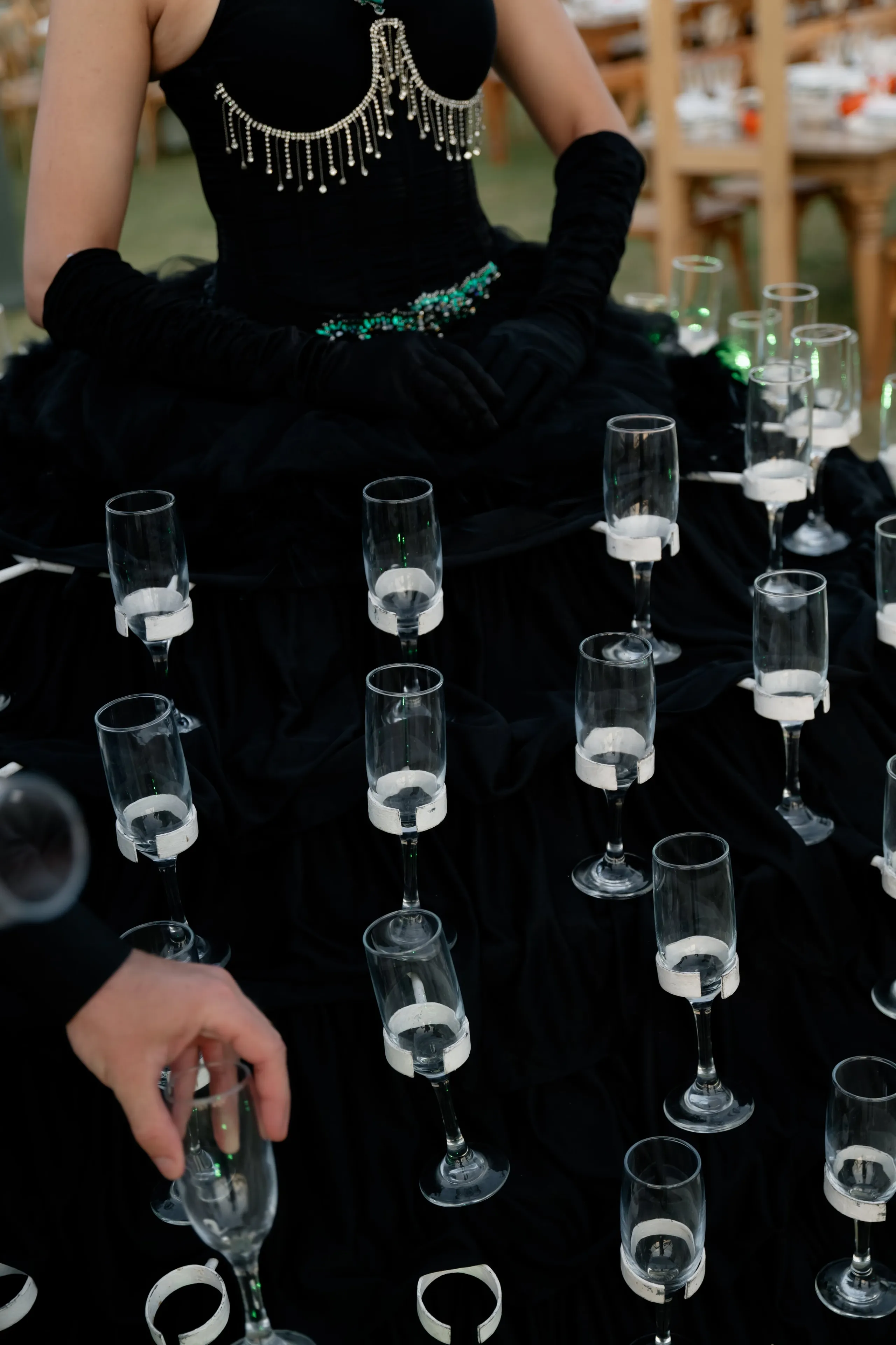 A woman in a black gown with a beaded fringe across the bodice and a green-jewelled waist, wearing long black gloves, stands among rows of empty champagne flutes fixed by white bands to a black fabric backdrop, with a hand reaching in from the left to hold one. Behind her, wooden chairs and set tables are visible. This is a wedding and event styling setup at Crete Golf Club in Chersonisos, Greece.
