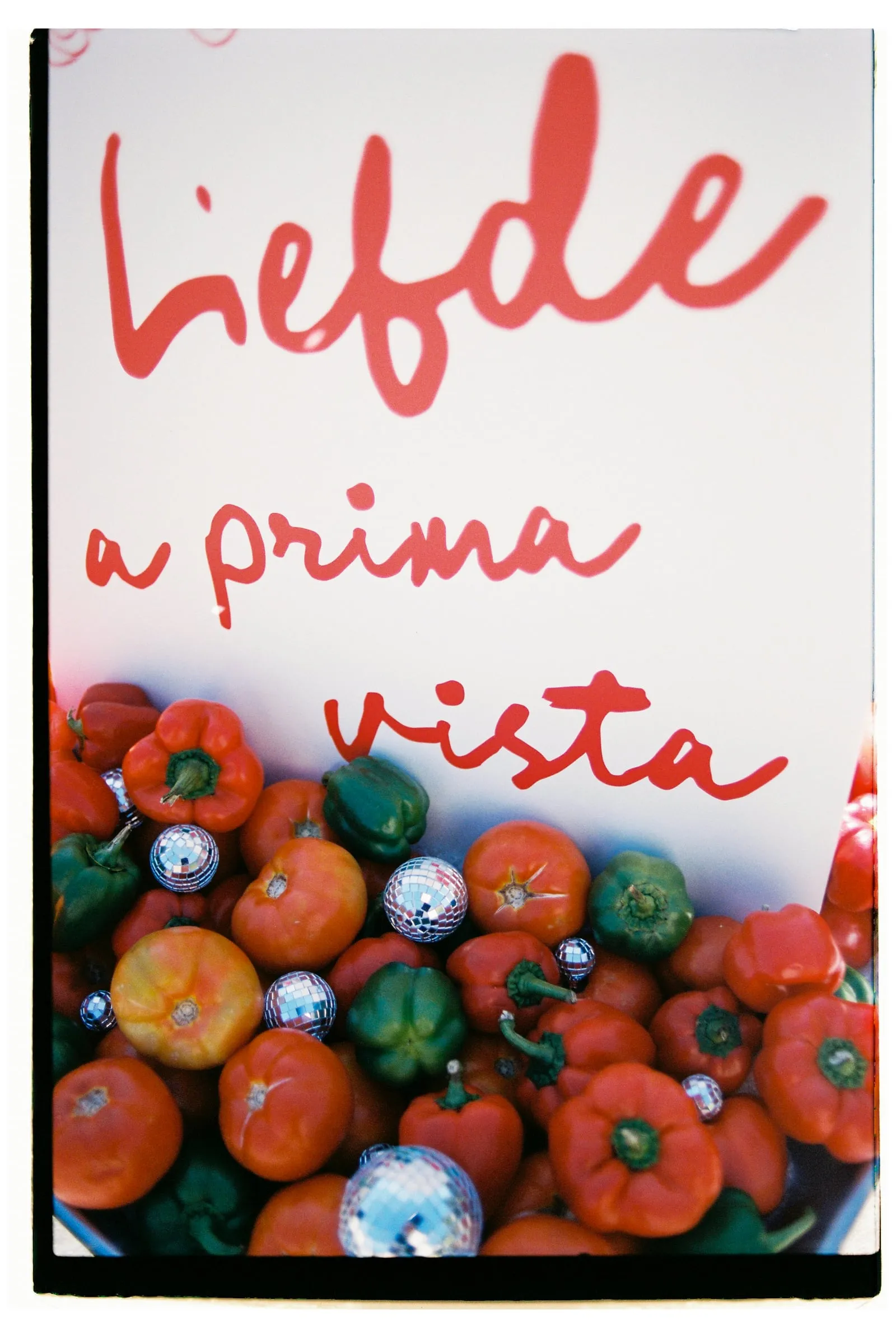 A close-up of a bowl heaped with red and green peppers, tomatoes and small silver mirror balls, set against a white backdrop with the hand-painted red words "Liefde a prima vista" (love at first sight) in loose brushstrokes. The playful mix of produce and sparkle, shot on film, sets a cheerful, romantic tone for a wedding story from Wine Dark Sea Villas in Rodia, Crete, Greece.