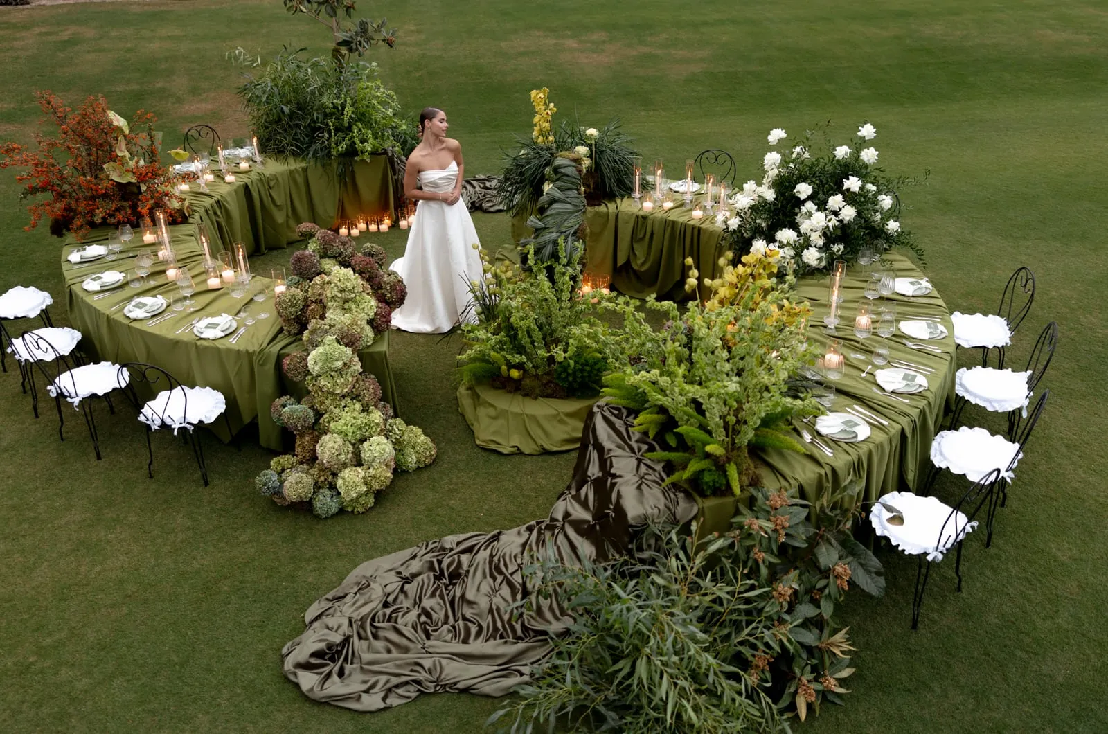 A bride in a strapless white satin gown stands on a lawn between curved tables draped in olive-green cloth, set for a wedding dinner at Ktima Roxani Estate in Armenoi, Crete, Greece. Arrangements of white roses, green hydrangeas fading to mauve, trailing eucalyptus, ferns and branches of orange-red berries surround the tables, while a dark olive satin train pools across the grass and white chairs line the edges. Taken from above, the scene is soft and green, with flickering candles along the tables.