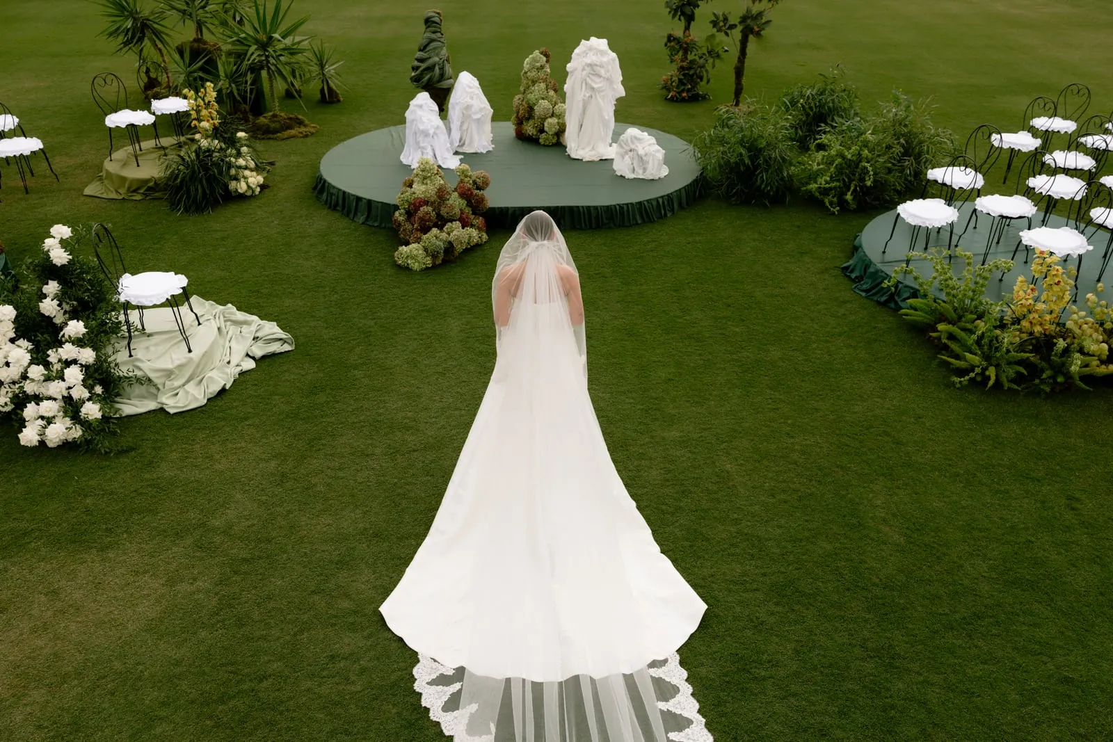 A bride seen from behind, her long lace-edged veil and train spilling across the lawn, walks toward a circular green-draped stage at Ktima Roxani Roxani Estate in Crete, Greece, where white and blush draped forms stand beside sculpted green foliage. Round stages to either side hold white-cushioned chairs, and clusters of white roses, hydrangeas and yellow-green orchids sit among palms and ferns on the vivid green grass. The aerial view and soft, overcast light give the wedding ceremony a calm, dreamlike, nature-inspired mood.