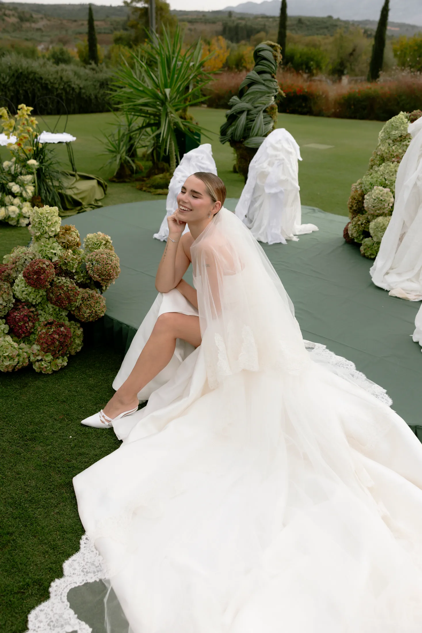 A bride in a flowing ivory gown and sheer lace-edged veil sits on the edge of a green circular platform, laughing with her chin resting on one hand. Beside her, clusters of green and burgundy hydrangeas spill across the lawn, with white-draped chairs and tall palms behind her. This is a wedding ceremony set-up at Ktima Roxani Estate in Crete, Greece, with soft daylight and rolling hills in the distance.