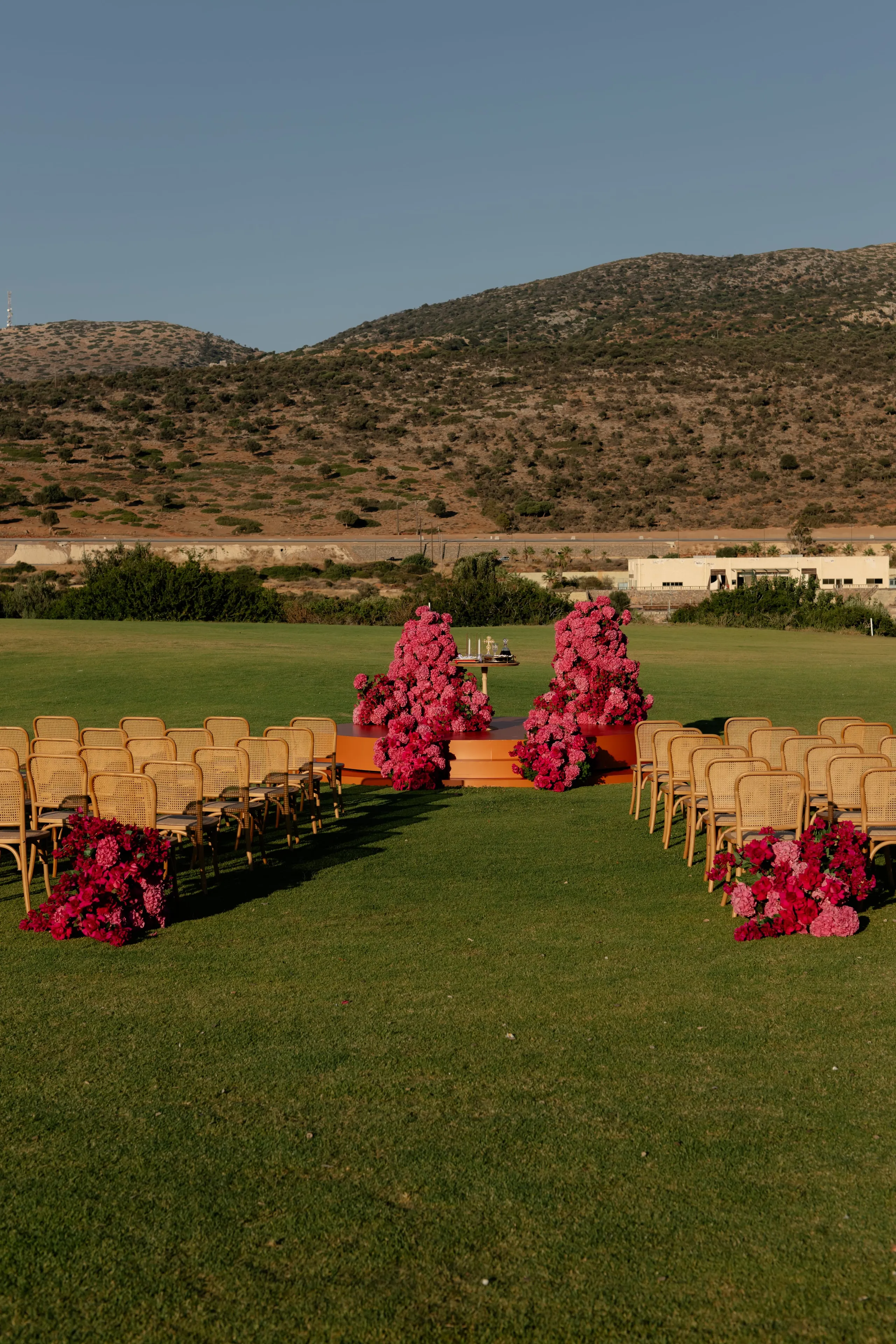 Rows of woven cane chairs frame a grassy aisle leading to a raised orange platform, where two tall, sculptural clusters of pink and magenta flowers rise on either side of a small round table. Low floral arrangements in the same hot pinks sit at the ends of the rows, and dry, scrubby hills rest under a clear evening sky. This wedding ceremony flower design sits on the lawn of Crete Golf Club in Chersonisos, Greece.