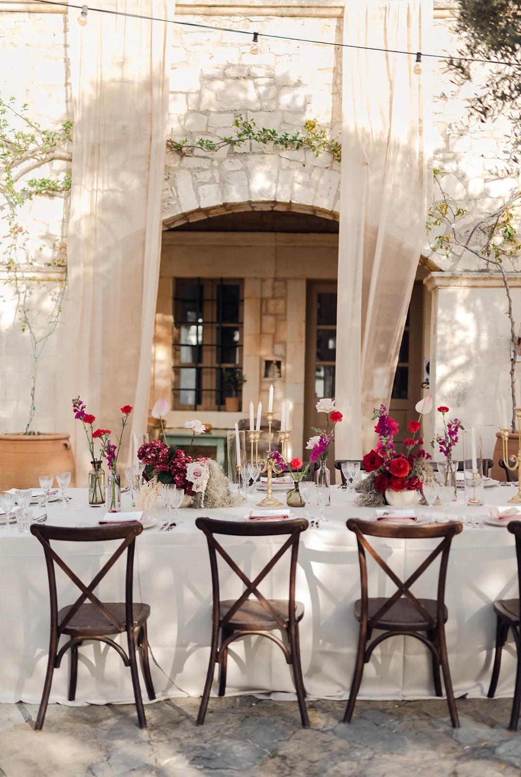 A long wedding table dressed in a white cloth, set outdoors at Agreco Farm in Greece, with dark wooden cross-back chairs lined up along one side. Low centrepieces of deep burgundy hydrangea, pale pink roses, fuchsia stock, red ranunculus and red carnations sit in small glass bottles and vases, interspersed with tall gold candlesticks and white taper candles. The warm, dappled light and soft cream drapes behind the table give it a relaxed, romantic feel.