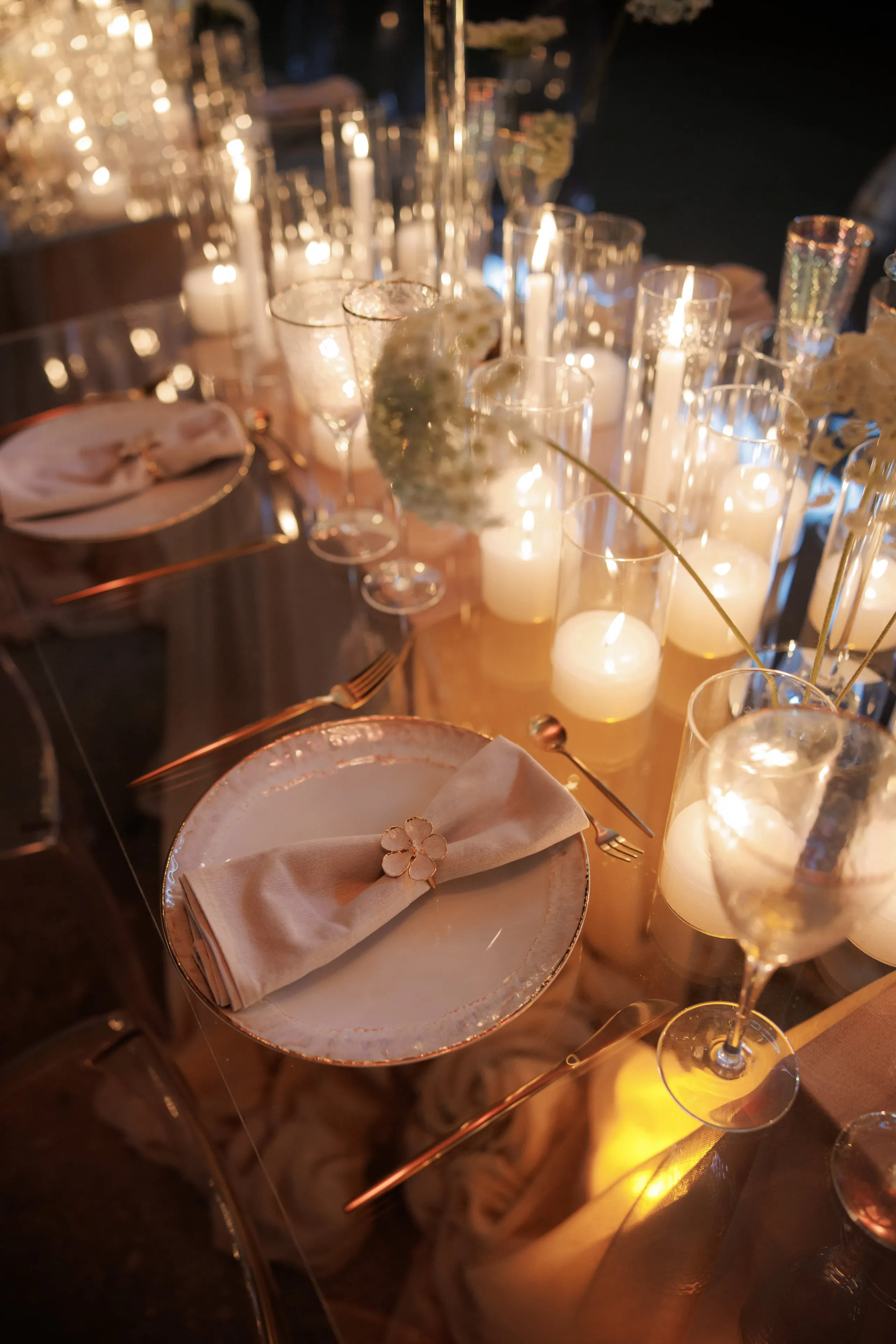 A candlelit wedding table in Rethimno, Crete, Greece, set on a glass-topped table with a blush linen napkin folded on a cream plate rimmed in copper, its ring a small golden flower. Tall glass hurricane candle holders glow amber along the table, and sprigs of pale, feathery white blooms rise from slim vases among the stemware. The warm light and soft reflections create an intimate, elegant evening mood.