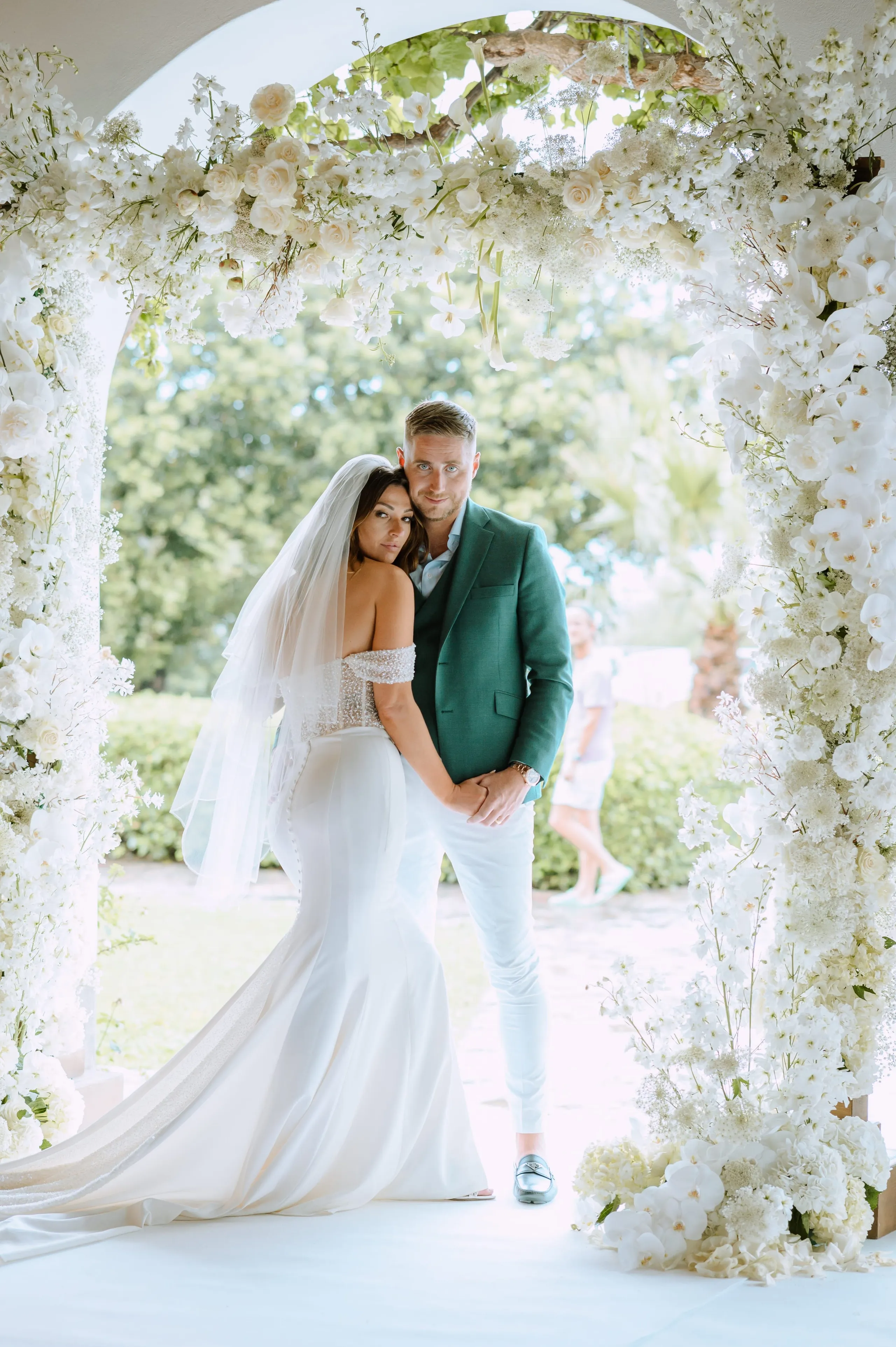 A bride and groom stand close together under a tall white floral arch at Minos Beach Art Hotel in Agios Nikolaos, Crete, Greece. The arch is dense with white roses, white orchids, delphinium-like spires and lacy white filler flowers, with trailing blooms framing the couple on both sides. She wears an off-shoulder fitted gown with a long train and a soft veil, and leans her head against his shoulder as he holds her hand; he wears a deep green blazer with pale trousers. Behind them, soft green trees and a blurred figure walking across a garden path give a bright, airy, summery feel.