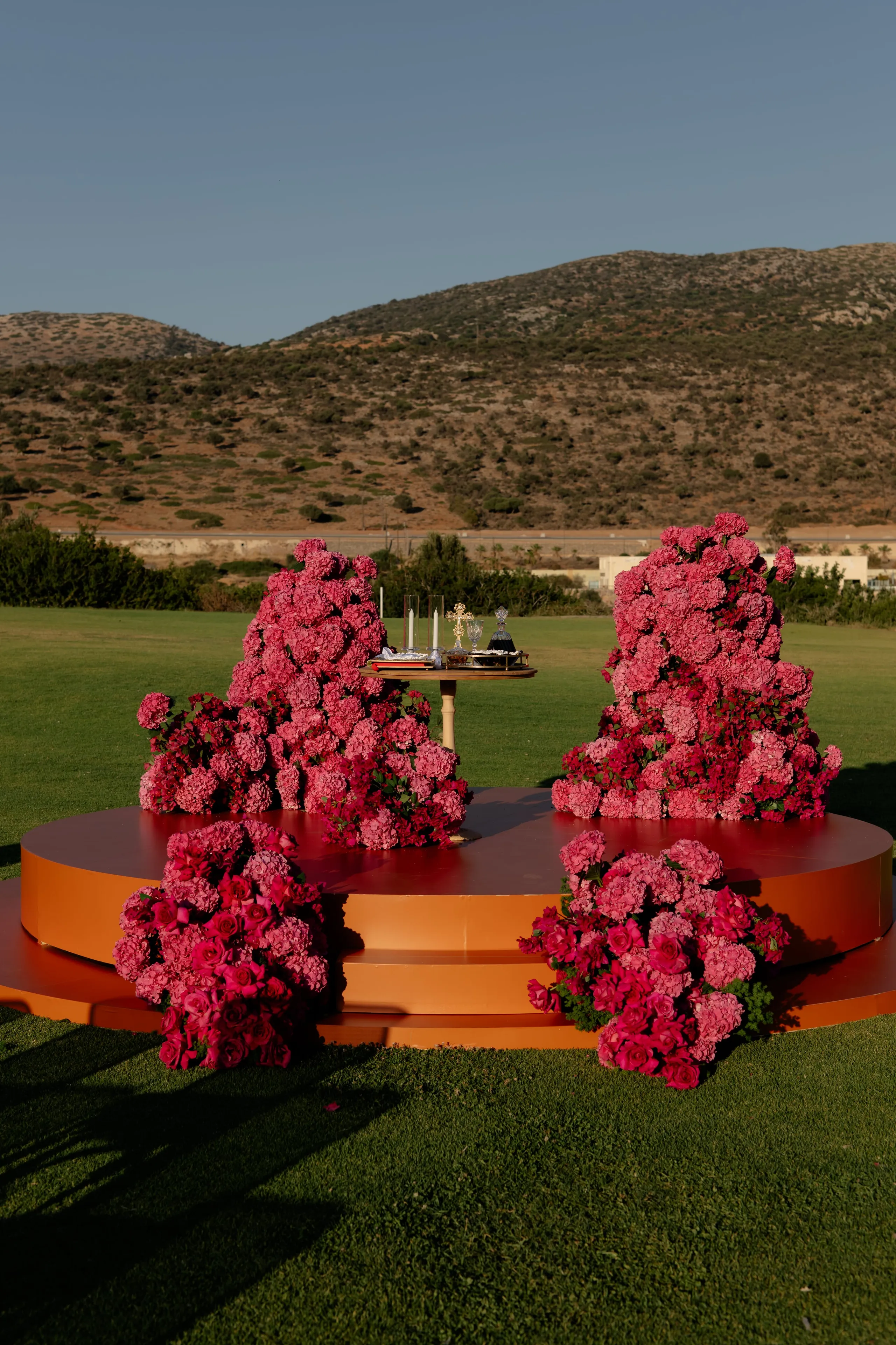 A circular orange-toned stage set on a green lawn at Crete Golf Club in Chersonisos, Greece, holds four dense clusters of pink and deep rose flowers, most likely hydrangeas and garden roses. Two tall mounds rise at the back on either side of a small gold table bearing candles and ceremonial items, while two lower clusters sit at the front corners. Hills dry and golden in the late light rise behind, under a clear blue sky, making this a sculptural wedding ceremony setting in Greece.