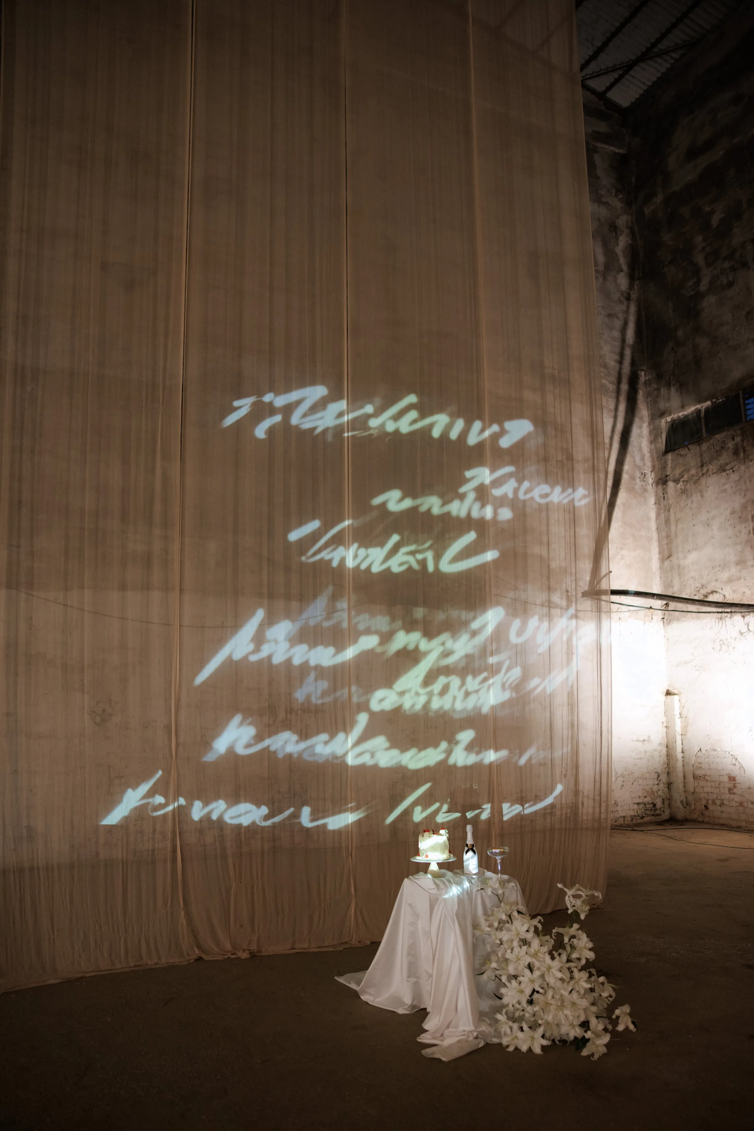 A large sheer cream curtain fills the tall, weathered concrete hall, with glowing handwritten script projected across its folds. In the lower right, a small white-draped table holds a tiered cake, a champagne bottle and a glass, with a cascade of white lilies spilling across the floor beside it. This wedding flower display, from The Extravaganza Flower, is set in Rethimno, Crete, Greece, and the soft, moody light gives the scene a quiet, dreamlike feel.