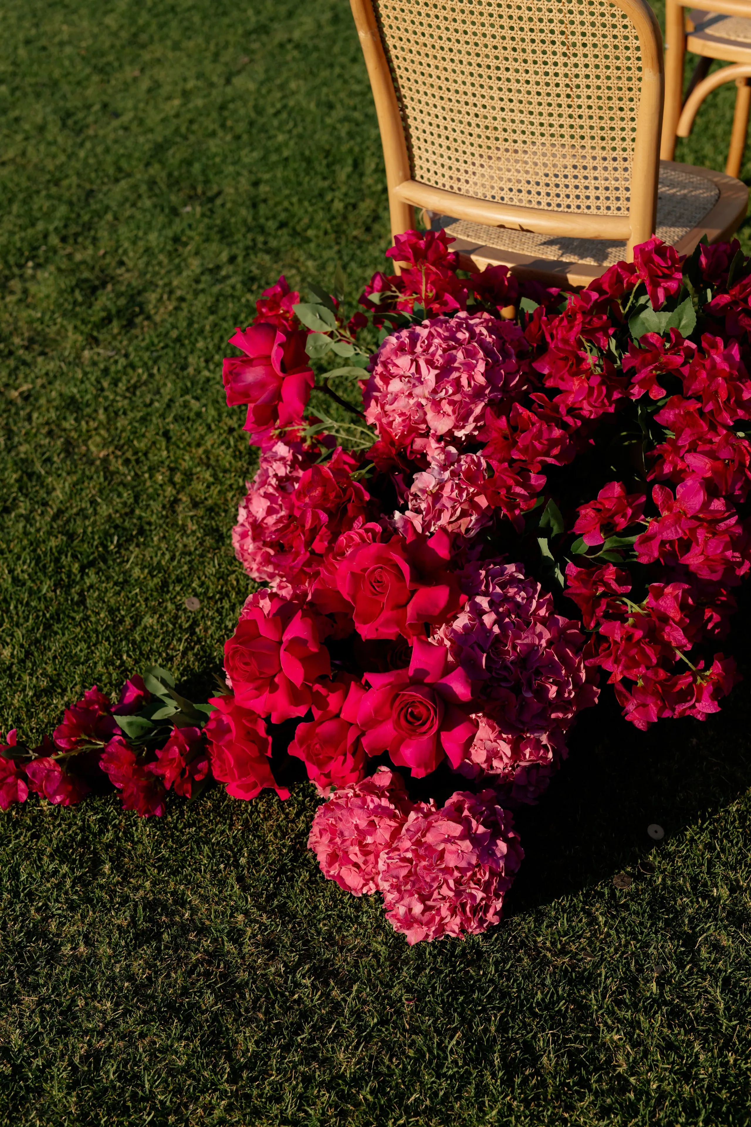 A cascade of hot pink and magenta flowers spills across green lawn at Crete Golf Club in Chersonisos, Greece, forming a sculptural ceremony installation for a wedding. Pink hydrangea heads and deep pink garden roses are woven with trailing bougainvillea and small green eucalyptus leaves. A woven cane chair sits behind the floral mass in soft late-afternoon sunlight, with long shadows across the grass.
