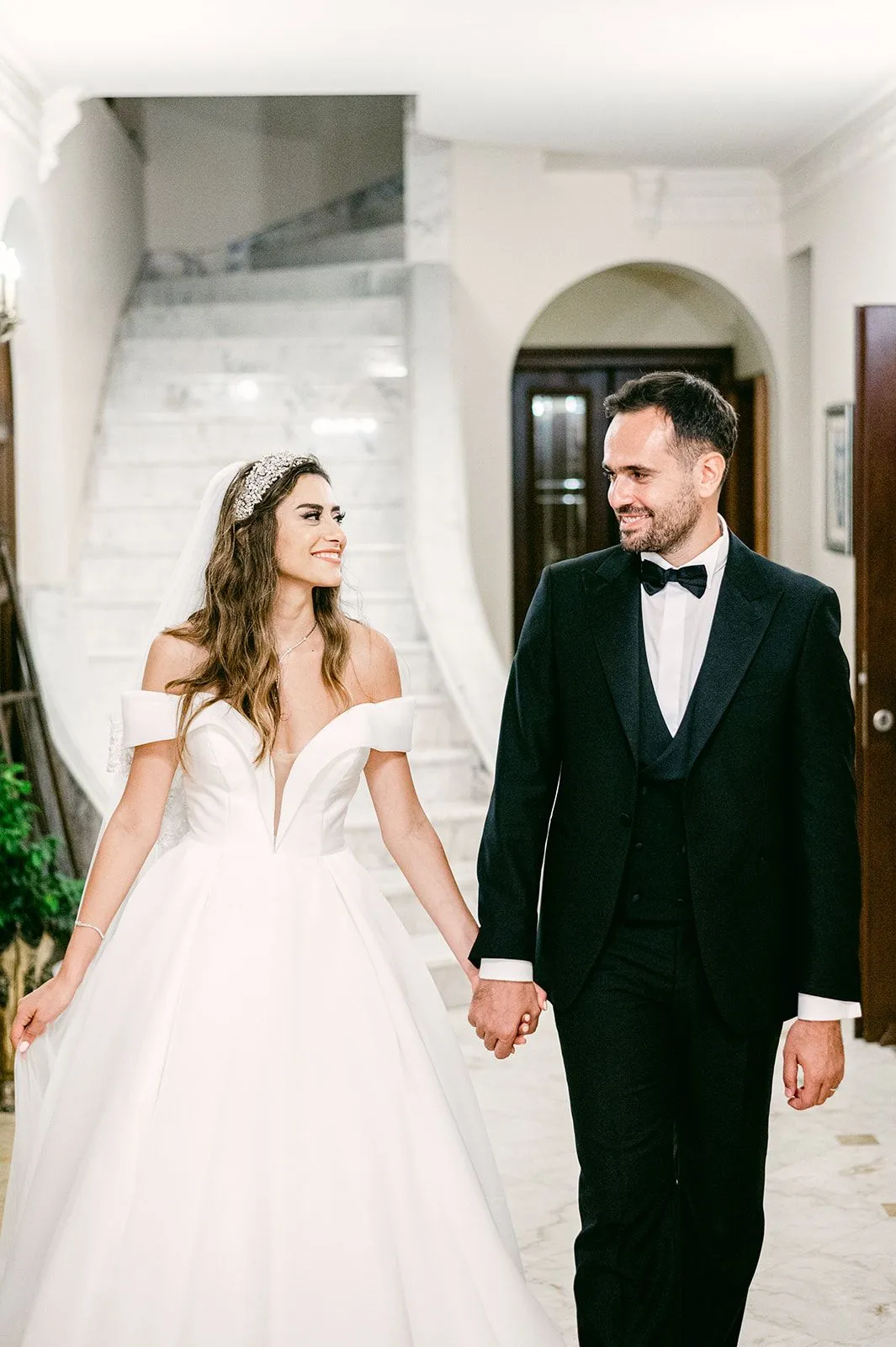 A bride and groom walk hand in hand through a bright marble hallway at Κτήμα Χατζή in Kerateas, Athens, Greece. She wears an off-the-shoulder white ballgown with a sweetheart neckline and a jewelled tiara in her wavy hair, and he wears a black tuxedo with a bow tie. Both are smiling and looking toward each other, with a marble staircase and an arched doorway behind them, a soft, elegant wedding-day moment.