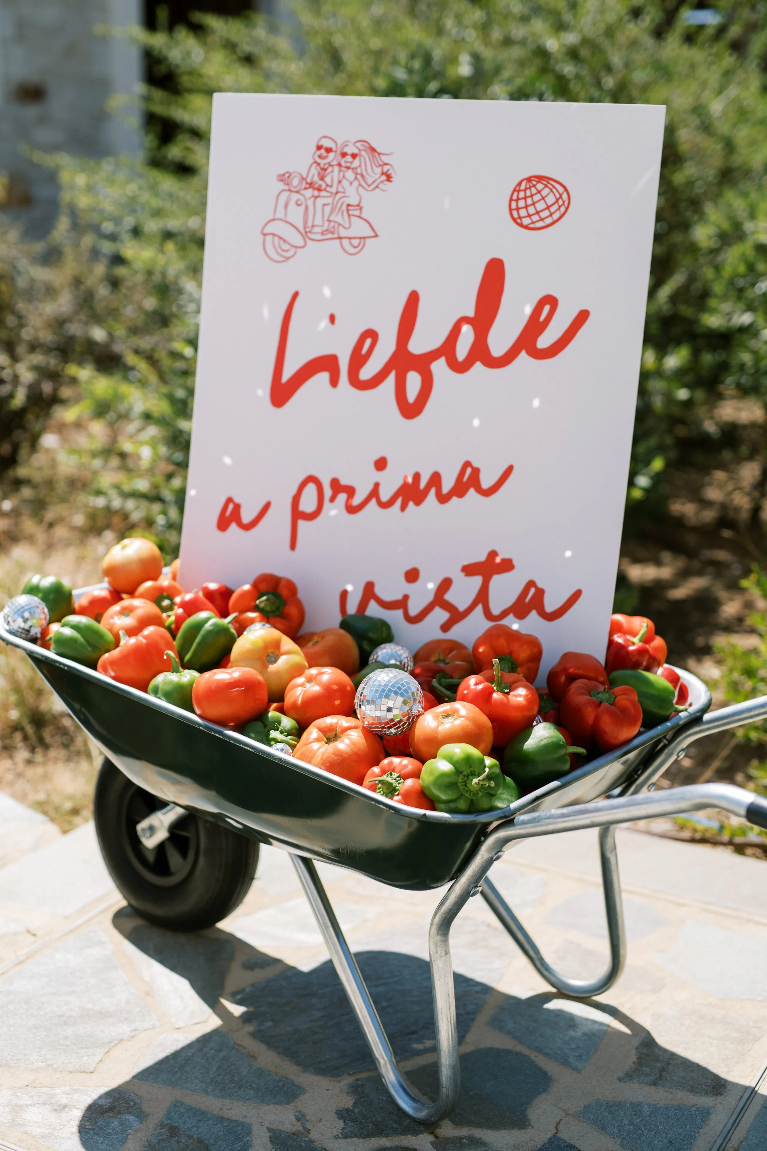 A black metal wheelbarrow on a stone terrace is heaped with ripe red and orange tomatoes, red and green peppers, and a few mirrored disco balls that catch the sunlight. Behind it stands a white sign with red hand-lettered script reading "Liefde a prima vista," decorated with a red line drawing of a couple riding a scooter. This playful wedding welcome display at Wine Dark Sea Villas in Rodia, Crete, Greece, is bright and cheerful, with green shrubs softly blurred behind it.