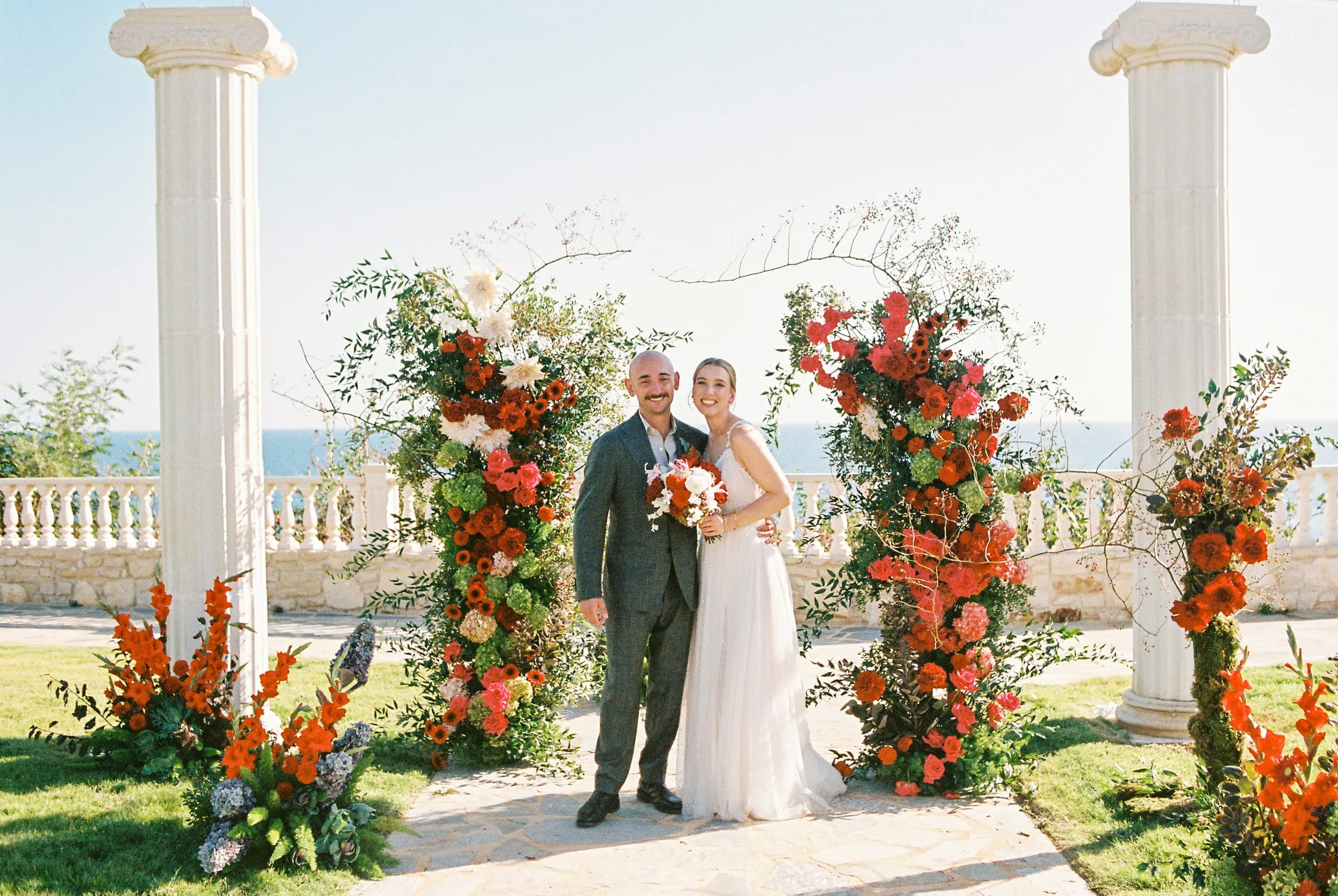 A bride and groom stand smiling together in front of a pair of tall flower arches of red and orange roses, dahlias and gladioli, with pink blooms, green hydrangeas and white dahlias woven through trailing branches. Two white Doric columns frame the scene, with a stone balustrade and the blue Mediterranean sea behind them under a pale sky. The wedding flowers, arranged for a garden ceremony at Wine Dark Sea Villas in Rodia, Crete, Greece, glow warmly in the sunlight.