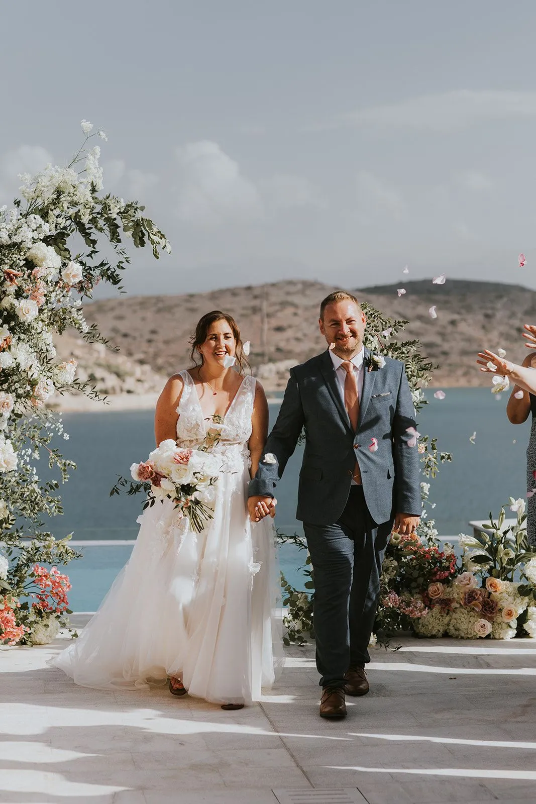 A bride and groom walk hand in hand down a stone terrace at Villa Gina in Schisma Eloundas, Crete, Greece, while petals drift through the air around them. Her lace-and-tulle gown is paired with a bouquet of white roses and blush blooms, and an arch of white flowers and trailing greenery frames the couple beside a calm blue sea and dry hills. He wears a grey suit with a dusty-pink tie, and both are smiling.