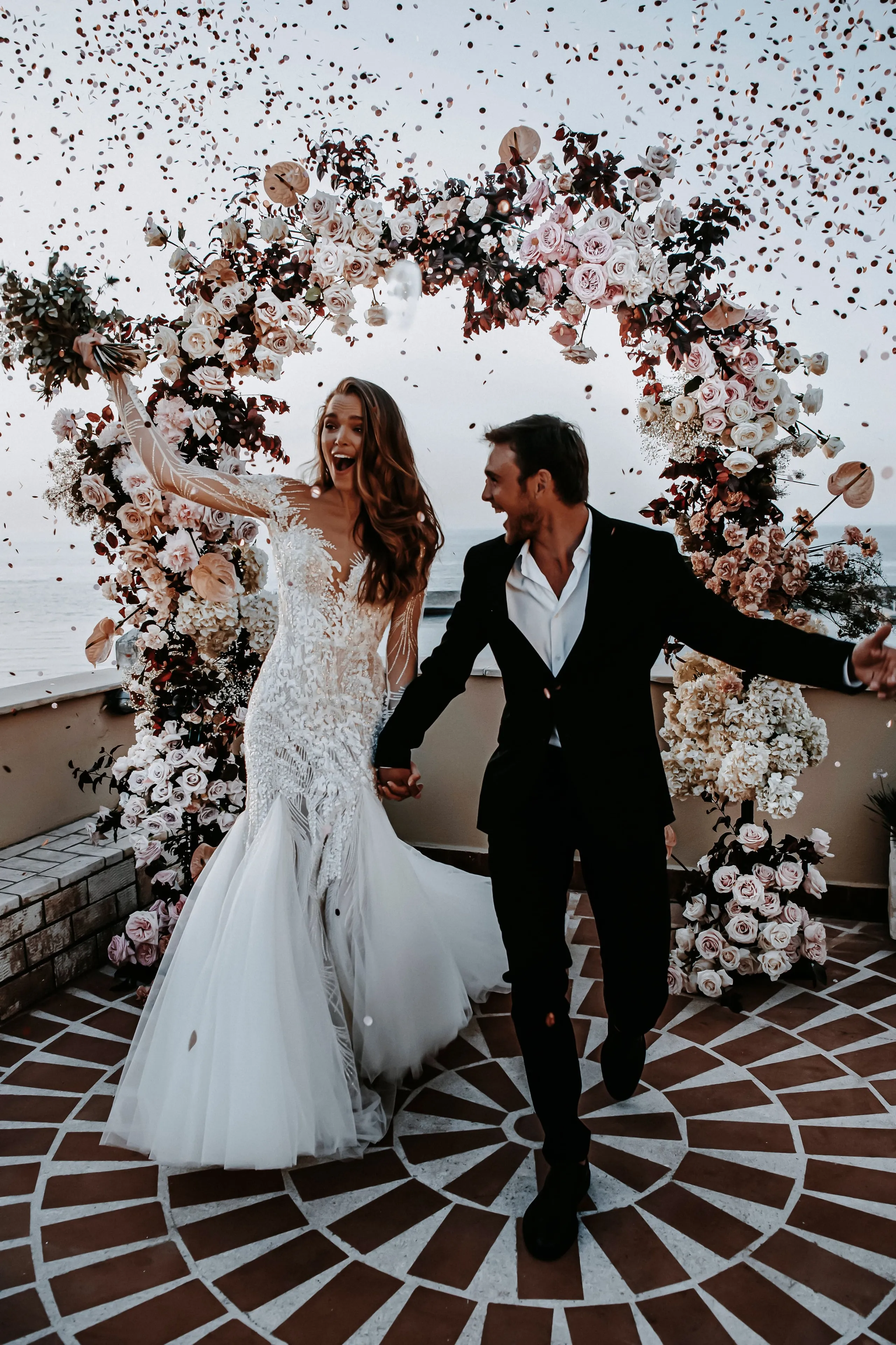 A joyful bride and groom step out from a tall floral arch of pale pink and cream roses, white hydrangeas and deep burgundy foliage, with confetti falling around them. She wears a fitted lace gown with sheer sleeves and raises one arm, while he, in a black suit and open white shirt, holds her hand and laughs. This wedding moment takes place on a rooftop terrace at Pandora Suites Hotel in Chania, Crete, Greece, with the sea behind them.