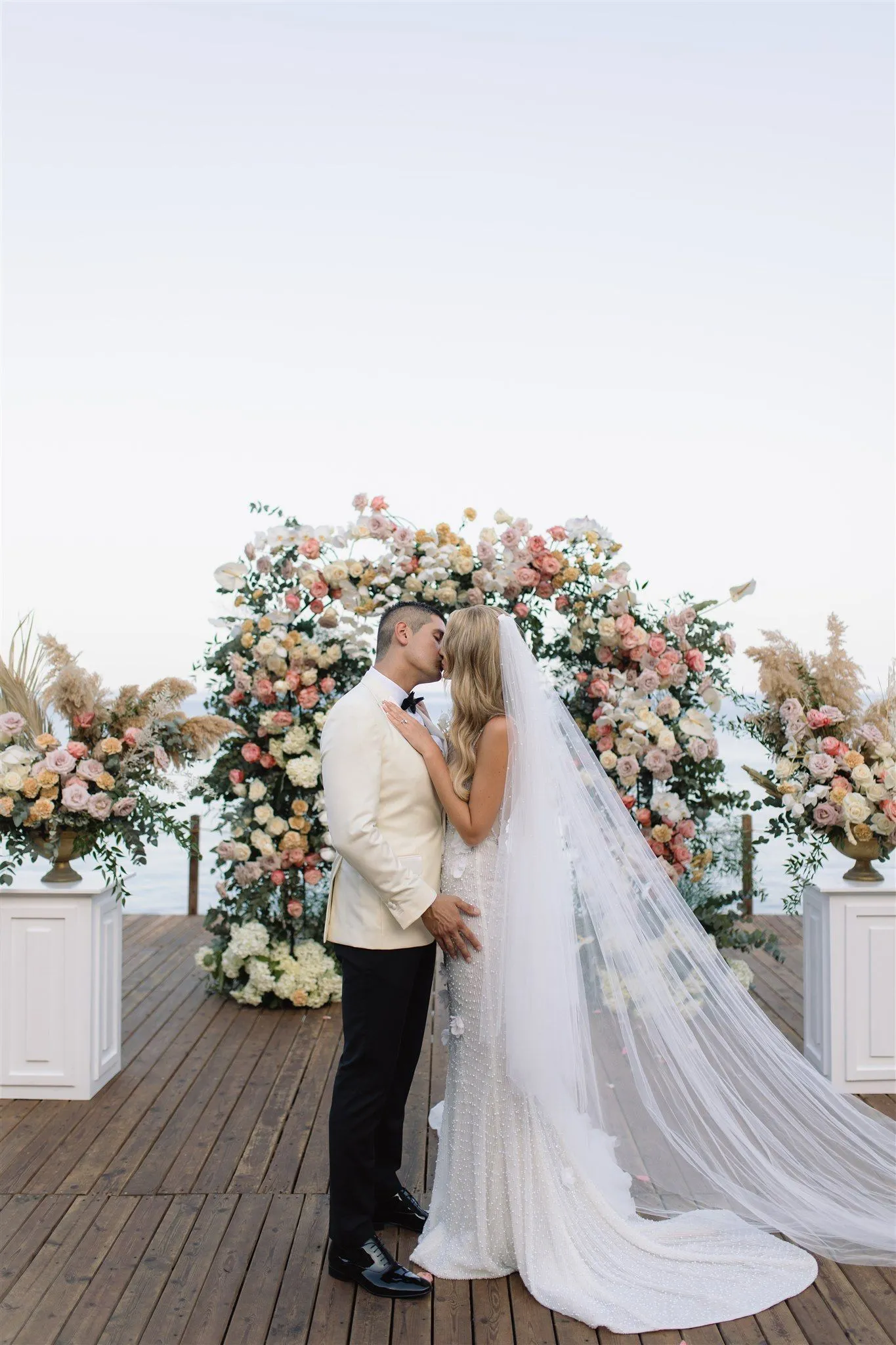 A bride and groom share a kiss in front of a full floral arch of blush, peach and cream roses with white hydrangeas and eucalyptus, flanked by tall urns of pink blooms and pampas grass, on a wooden deck at Lazarou Beach in Greece. The groom wears an ivory dinner jacket with a black bow tie and black trousers, while the bride's fitted, pearl-covered gown trails a long veil across the boards. The soft pastel palette and pale, open sky give the wedding flowers a calm, romantic glow.