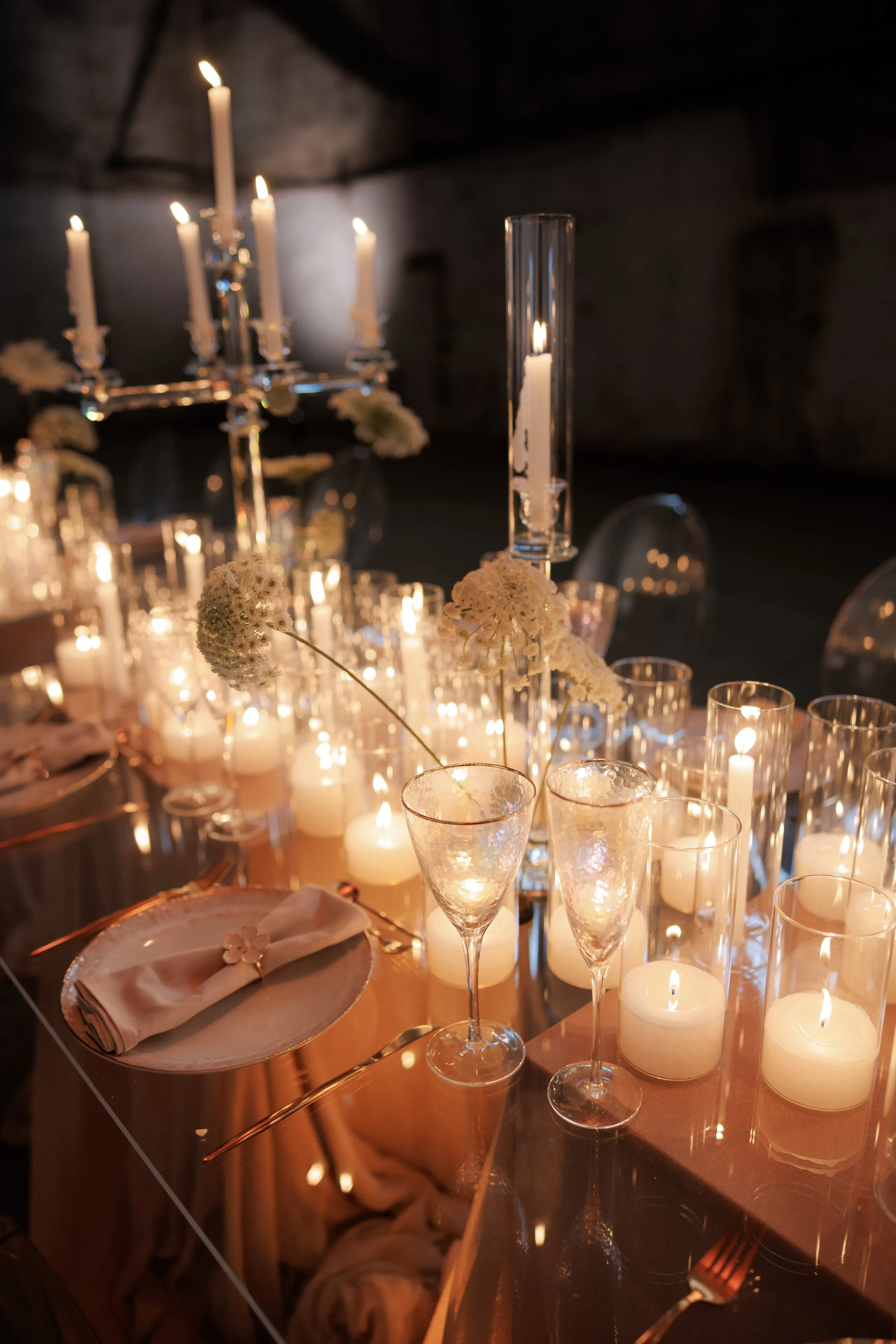 A candlelit wedding table in Rethimno, Crete, Greece, dressed in soft blush linen and a glass-topped surface that reflects the flames. Tall taper candles rise from a glass candelabra and a slender glass cylinder, while rows of pillar candles glow inside clear hurricane holders along the table. Pale cream allium blooms, with their round, lacy heads, are scattered among the glasses, and a folded blush napkin sits on a plate beside etched champagne flutes.