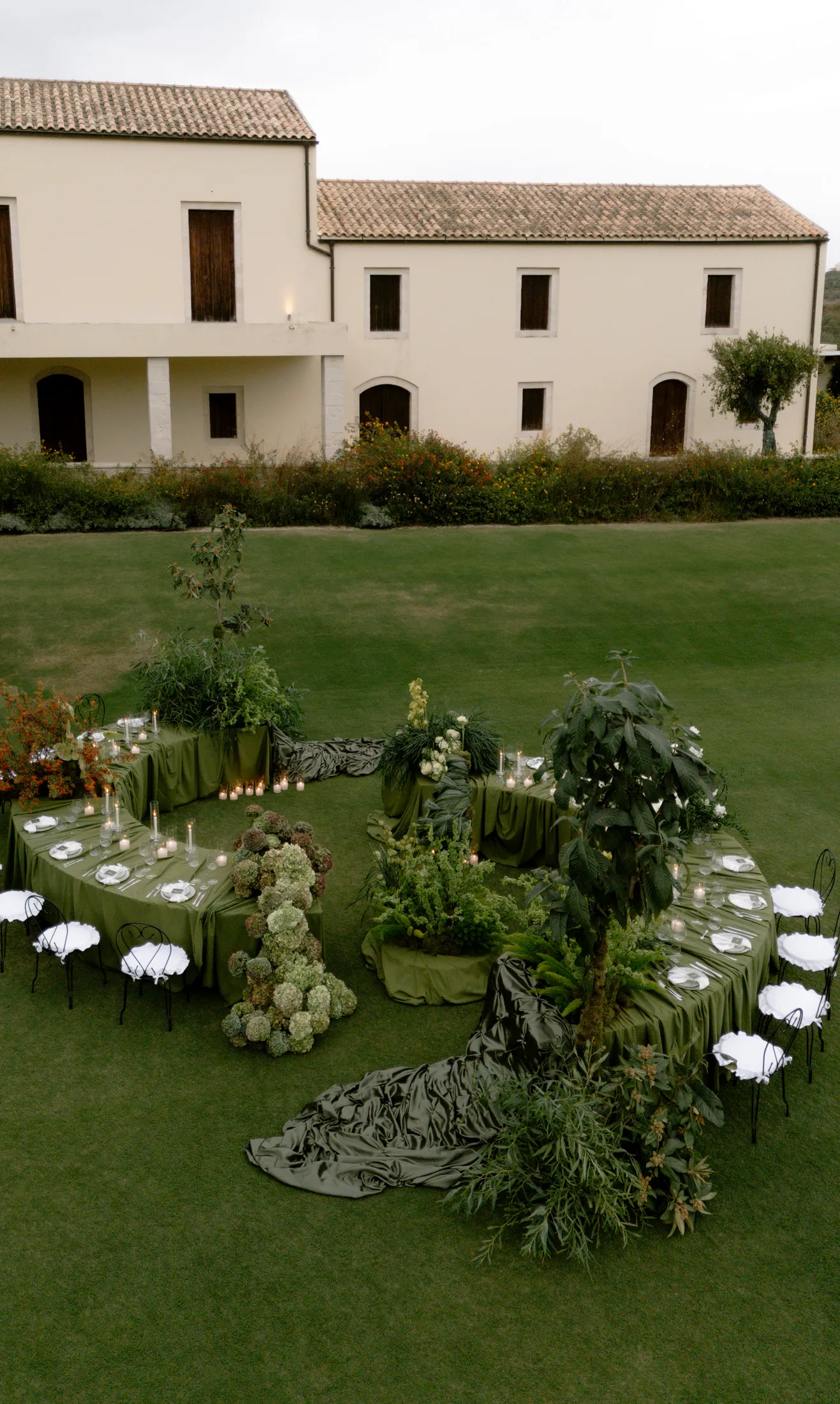 An aerial view of a wedding dining setup on the lawn of Ktima Roxani estate in Armenoi, Crete, Greece, with long tables draped in sage-green cloth arranged in a curving, open layout. Potted trees and lush greenery rise between the tables, with clusters of pale green hydrangeas and white blooms as centrepieces, and white wrought-iron chairs line the edges. Low tealight candles glow along the tables beneath a cream two-storey villa with terracotta roof tiles.