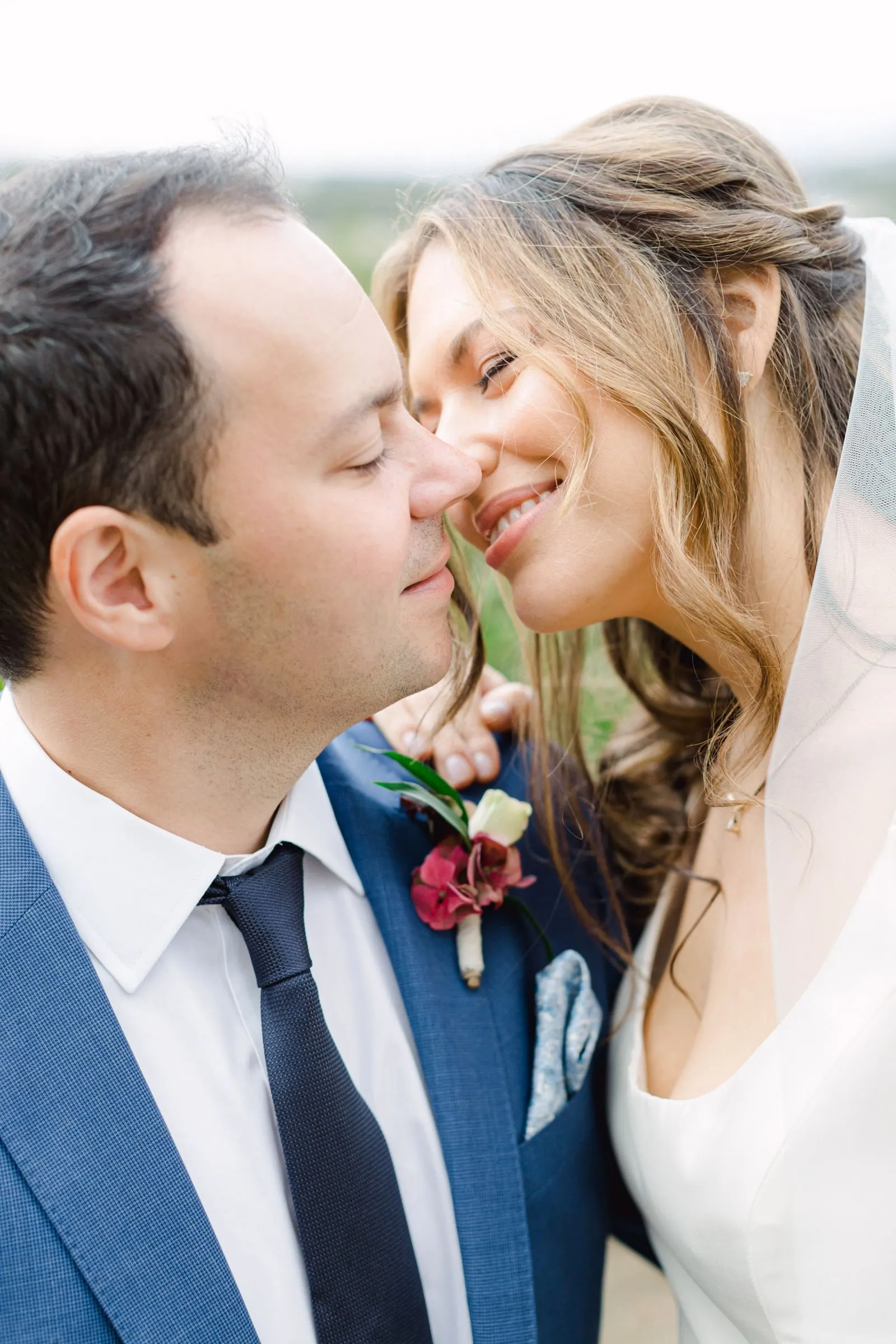 A close, tender portrait of a bride and groom at AgrecoFarms in Adele, Crete, Greece, their noses almost touching as they share a smile. The groom, on the left with his eyes closed, wears a navy suit, white shirt and dark tie, with a small boutonniere of deep pink blooms and a white rose pinned to his lapel. The bride, her wavy light-brown hair swept back and a sheer veil drifting at the edge of the frame, leans in with a soft smile, a green blur of countryside behind them.
