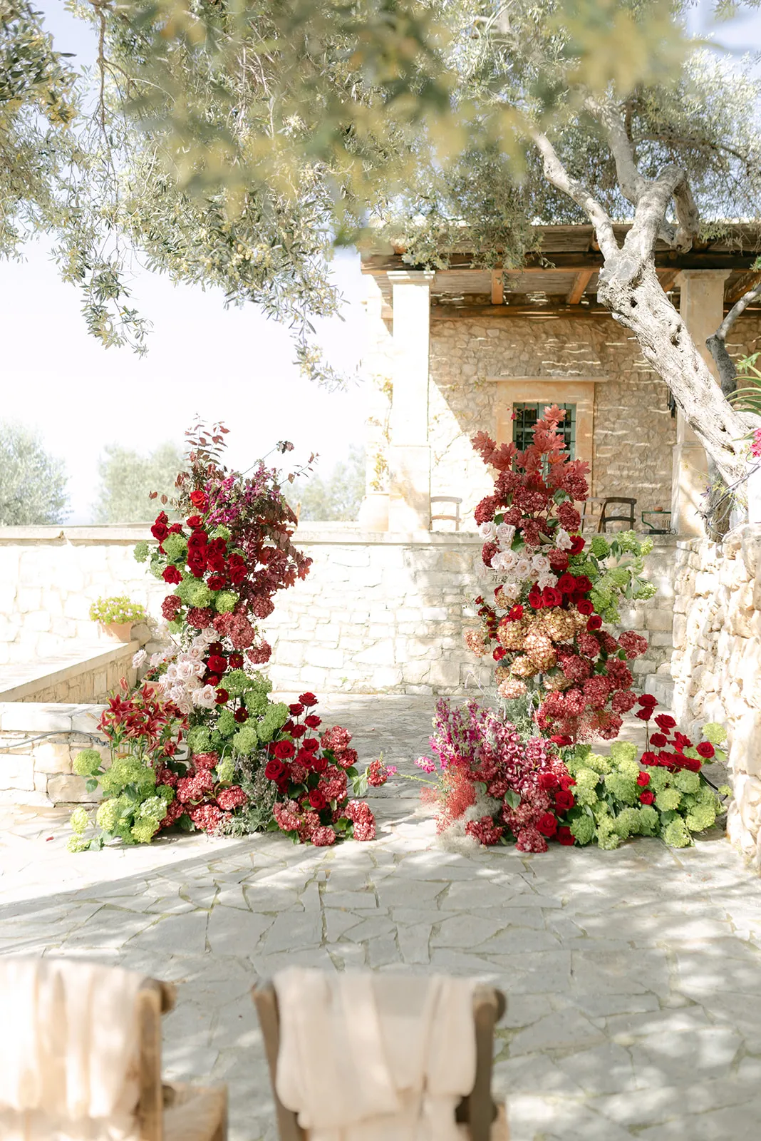 Two tall, cascading floral installations in deep red, blush and cream frame a stone-paved courtyard, with red roses, pink and burgundy hydrangeas, white roses and green viburnum spilling down from their tops. Olive branches overhead and a weathered stone farmhouse behind them set the scene in this wedding flower display at Agreco Farm in Greece.