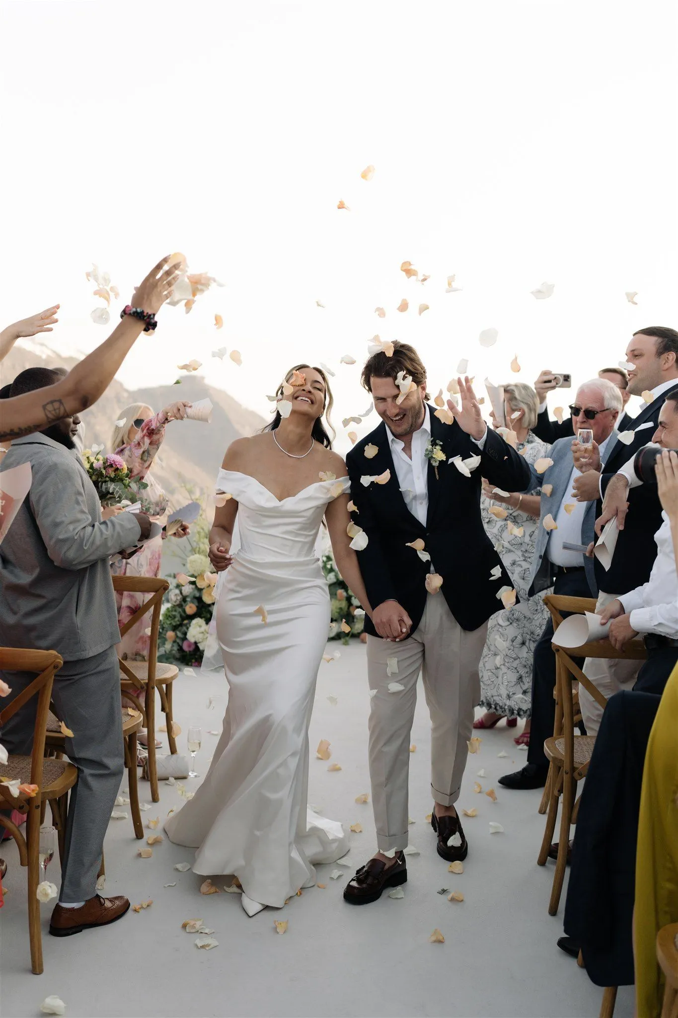 A bride in a fitted white satin gown with an off-the-shoulder neckline and a pearl necklace laughs as she walks down an aisle with her husband, who wears a navy blazer, open-necked white shirt and stone trousers. Guests on both sides throw pale peach and cream rose petals into the air above them, filling the bright, hazy sky. This is a wedding ceremony at Aenaon Villas in Santorini, Greece, with white-clothed cross-back chairs lining the aisle and garden flower arrangements framing the terrace.