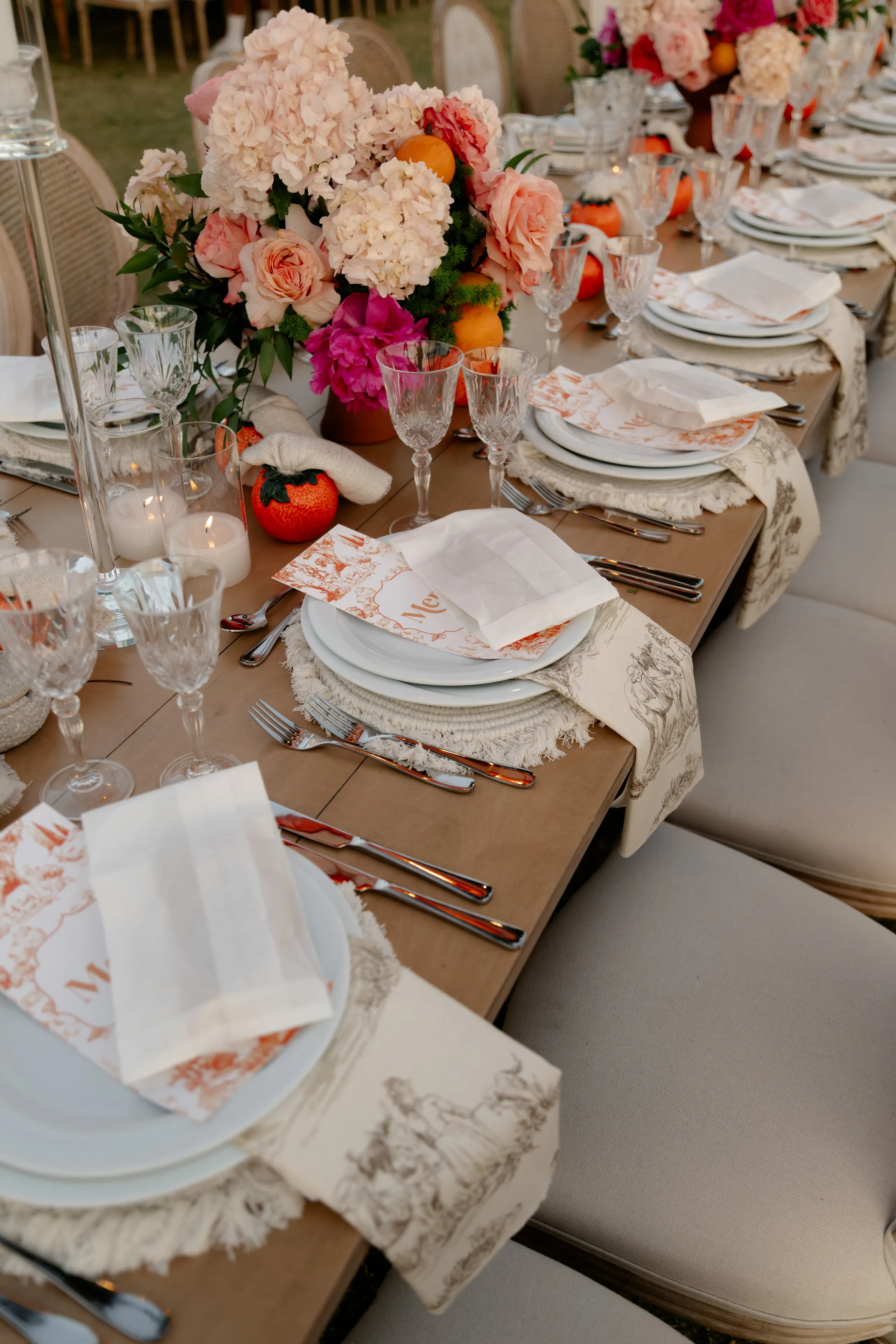 Wedding table flowers at Crete Golf Club in Chersonisos, Greece: a long wooden table set for a celebration, with a centrepiece of pale pink hydrangeas, peach garden roses and a hot pink bloom, tucked among orange fruit and green foliage. Each place has a white plate, folded white napkin, and a menu card and napkin edged in a toile-style print of orange-red and brown, with fringed cream placemats and clear wine glasses lining the table under soft evening light.