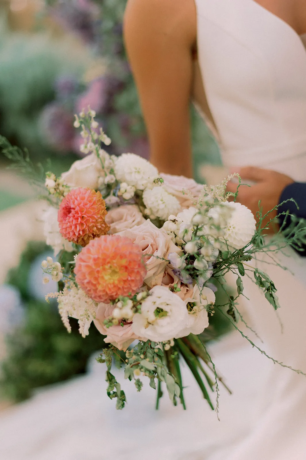 A loosely gathered bridal bouquet at Lake Vouliagmeni in Greece, with two coral-orange dahlias as its bold focal points. Around them sit blush garden roses, creamy white ranunculus, and white pompom dahlias, all softened by trailing green foliage, tiny white berries, and wispy astilbe plumes. The stems are bound in green, and the background is blurred into soft pale tones, giving a gentle, sunlit, romantic mood.