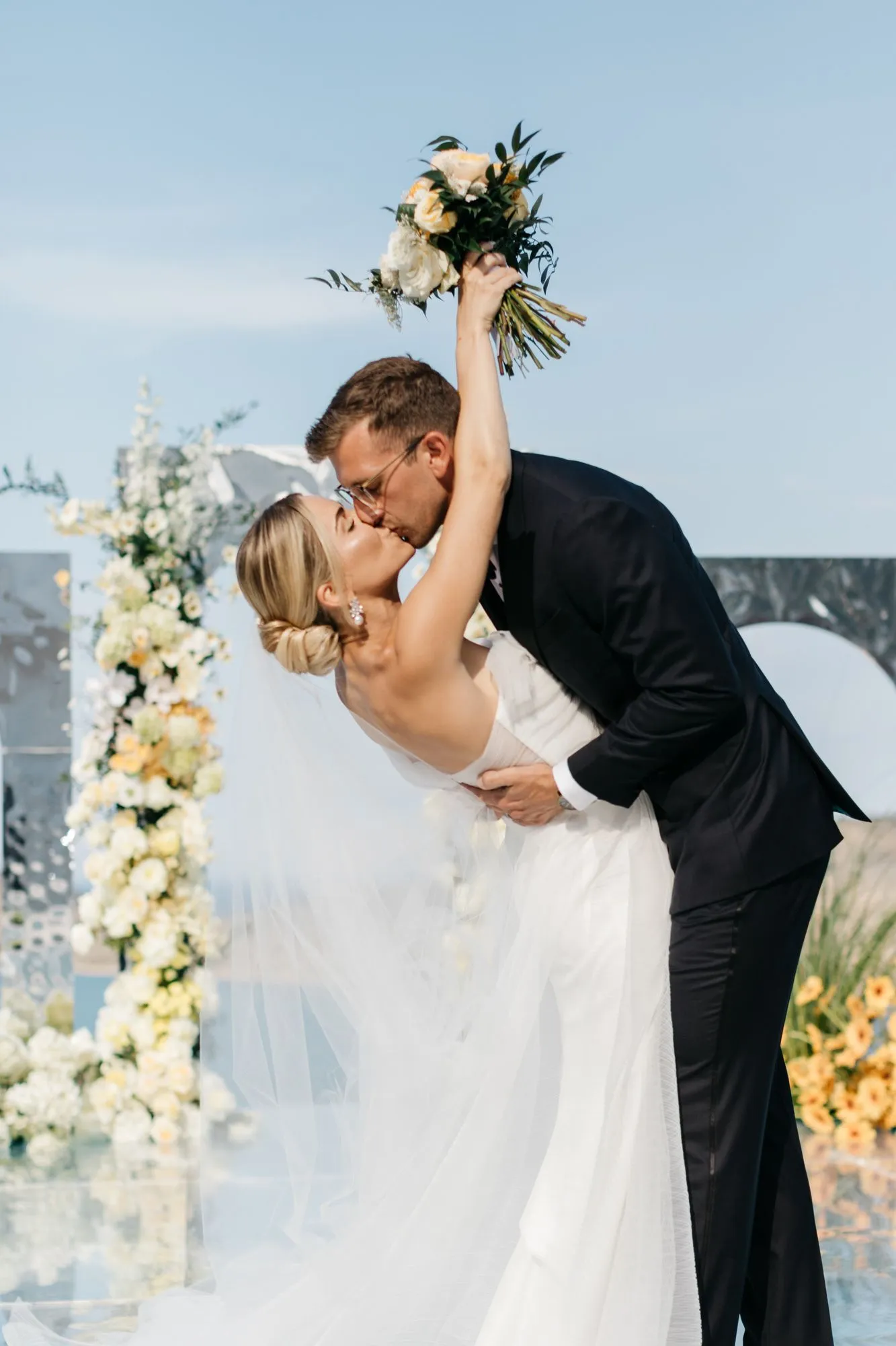 A groom in a dark suit and glasses dips his bride backwards as they kiss beneath a pale blue sky, her raised arm holding a bouquet of cream roses, white blooms and sprigs of green foliage. Behind them a tall column of white and pale yellow flowers rises beside a grey stone arch, part of the wedding ceremony decor at Dream Art Villa in Schisma Elountas, Crete, Greece. Her long white veil drifts in the breeze, and more yellow flowers sit along the glossy floor.
