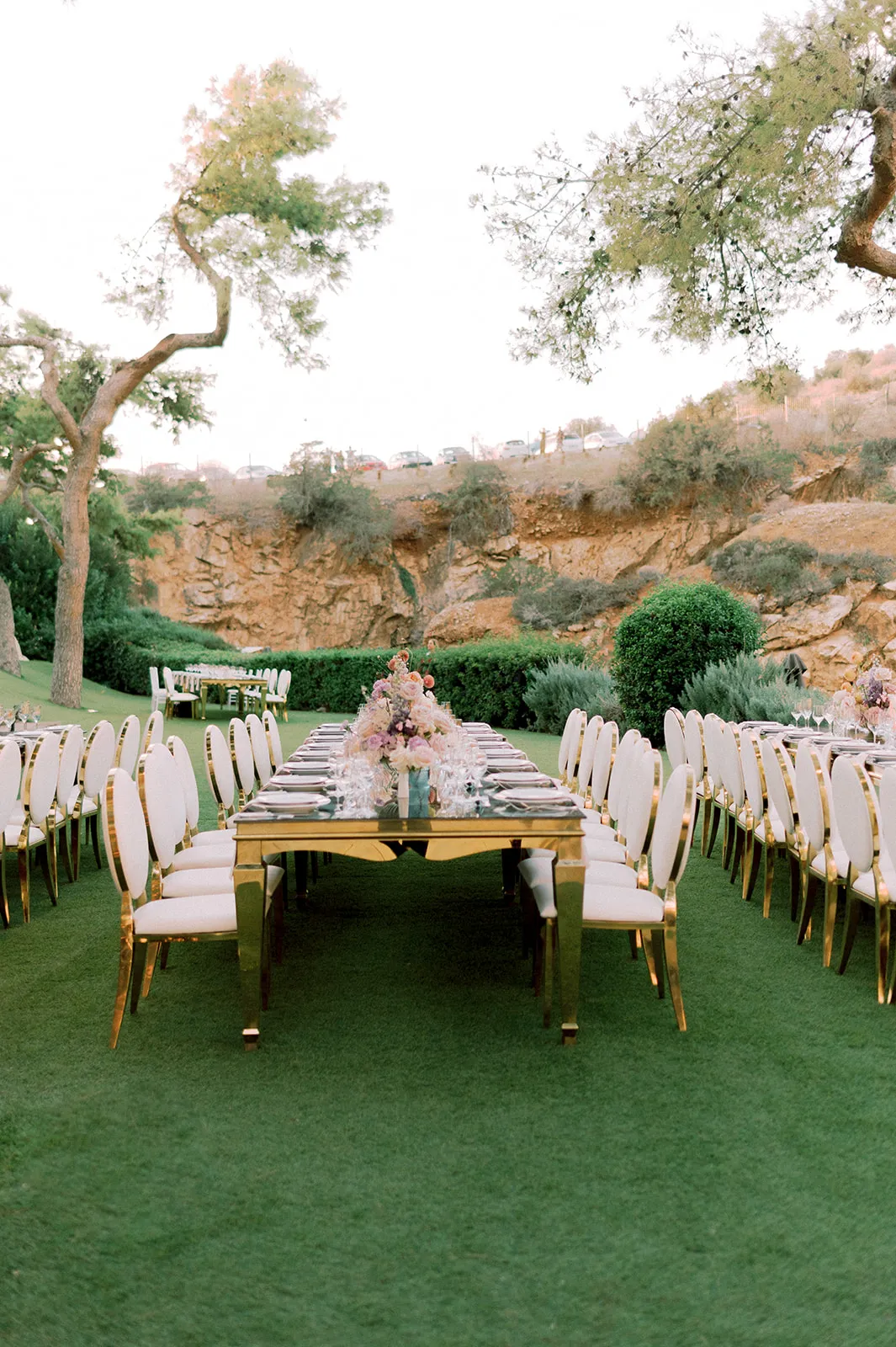 A long gold-framed banquet table runs across a green lawn, set with white plates and glassware and flanked by rows of white-cushioned chairs with gold trim. At its centre sits a tall arrangement of garden roses in blush, peach and lilac, with trailing foliage, for a wedding table in Lake Vouliagmeni, Greece. Olive trees, a rounded green shrub and a rocky cliff bathed in soft evening light frame the scene.