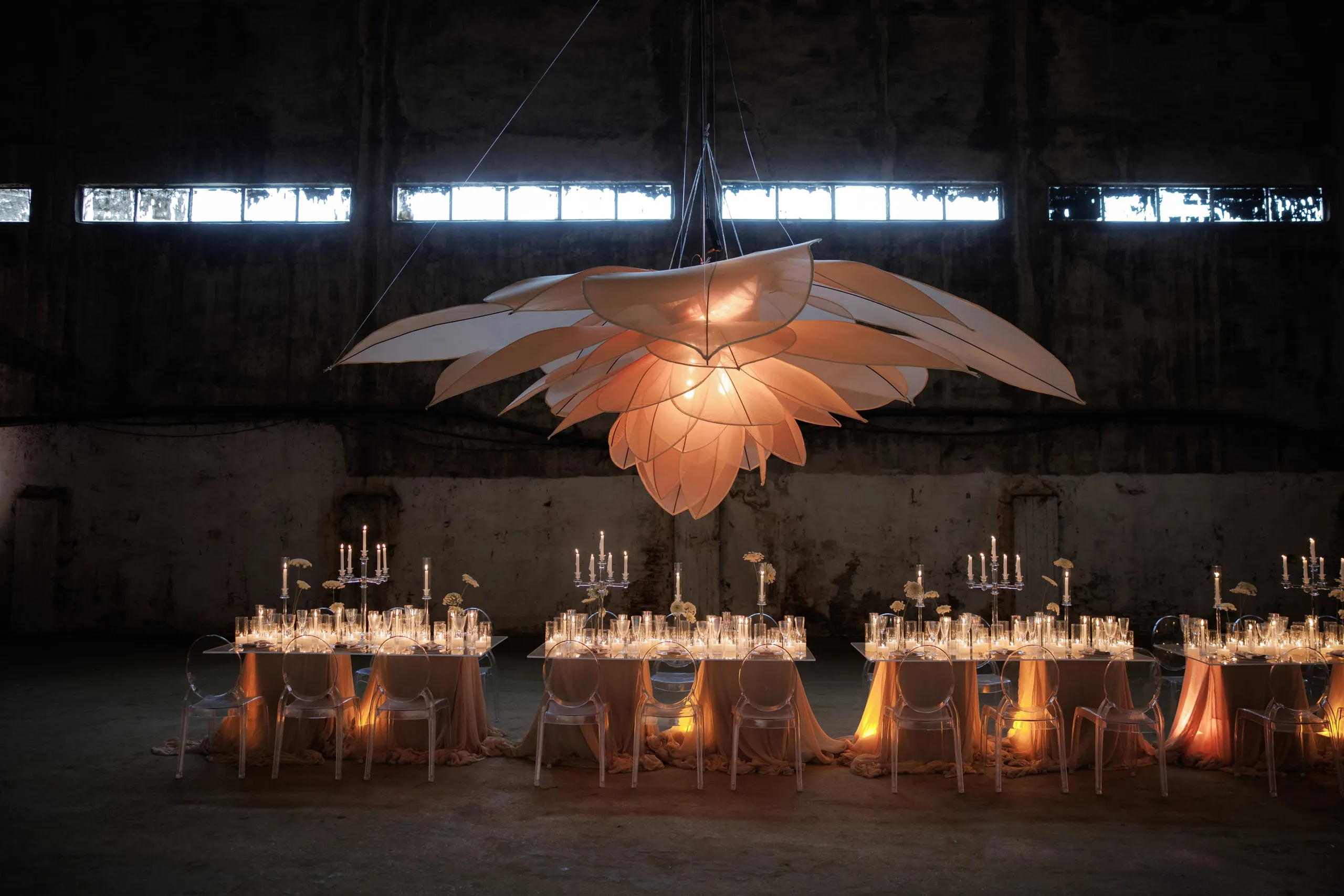 A vast, illuminated flower sculpture of layered peach petals hangs above three long tables in a dim, weathered industrial hall, with a soft glow spilling onto peach-draped tables, clear ghost chairs, and candelabra. Small cream blooms sit in low arrangements among the glassware. This is a wedding table setup in Rethimno, Crete, Greece, with the warm light and dark concrete walls creating a dramatic, intimate mood.
