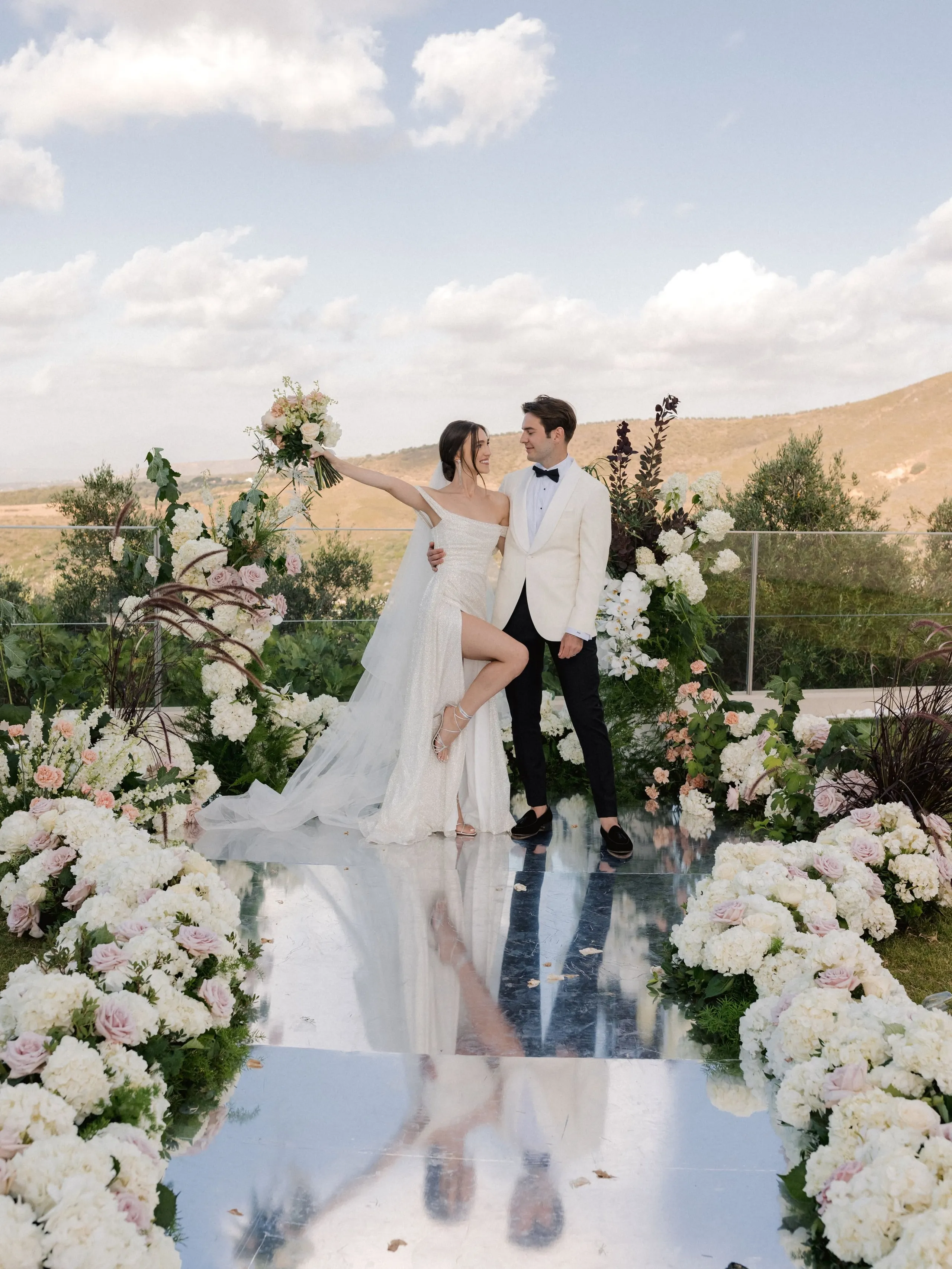 A bride in a sparkling one-shoulder gown lifts her bouquet high while lifting one leg to the side, as a groom in a cream dinner jacket and black bow tie smiles beside her. They stand on a mirror-like walkway lined with white hydrangeas and blush garden roses, with dark branches and tall grasses framing an arch behind them. Wedding flowers in Greece at Amphi Residence in Tria Monastiria, Crete, with hills and a soft blue sky beyond.