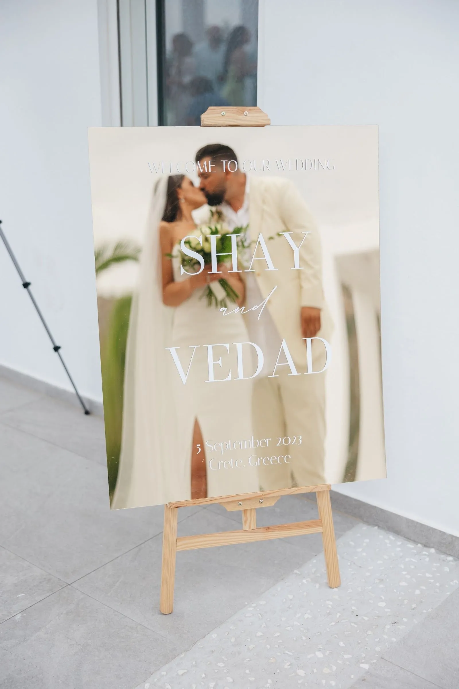 A large acrylic welcome sign on a wooden easel reads "Welcome to our wedding, Shay and Vedad, 5 September 2023, Crete, Greece," with a photo of the couple kissing printed beneath the white lettering. The bride wears a fitted white gown with a long veil and holds a bouquet of white blooms and green foliage, while the groom wears a cream suit. The sign stands on a pale grey terrazzo floor against a white wall at Villa Imperial in Crete, Greece, a soft, airy wedding welcome display.