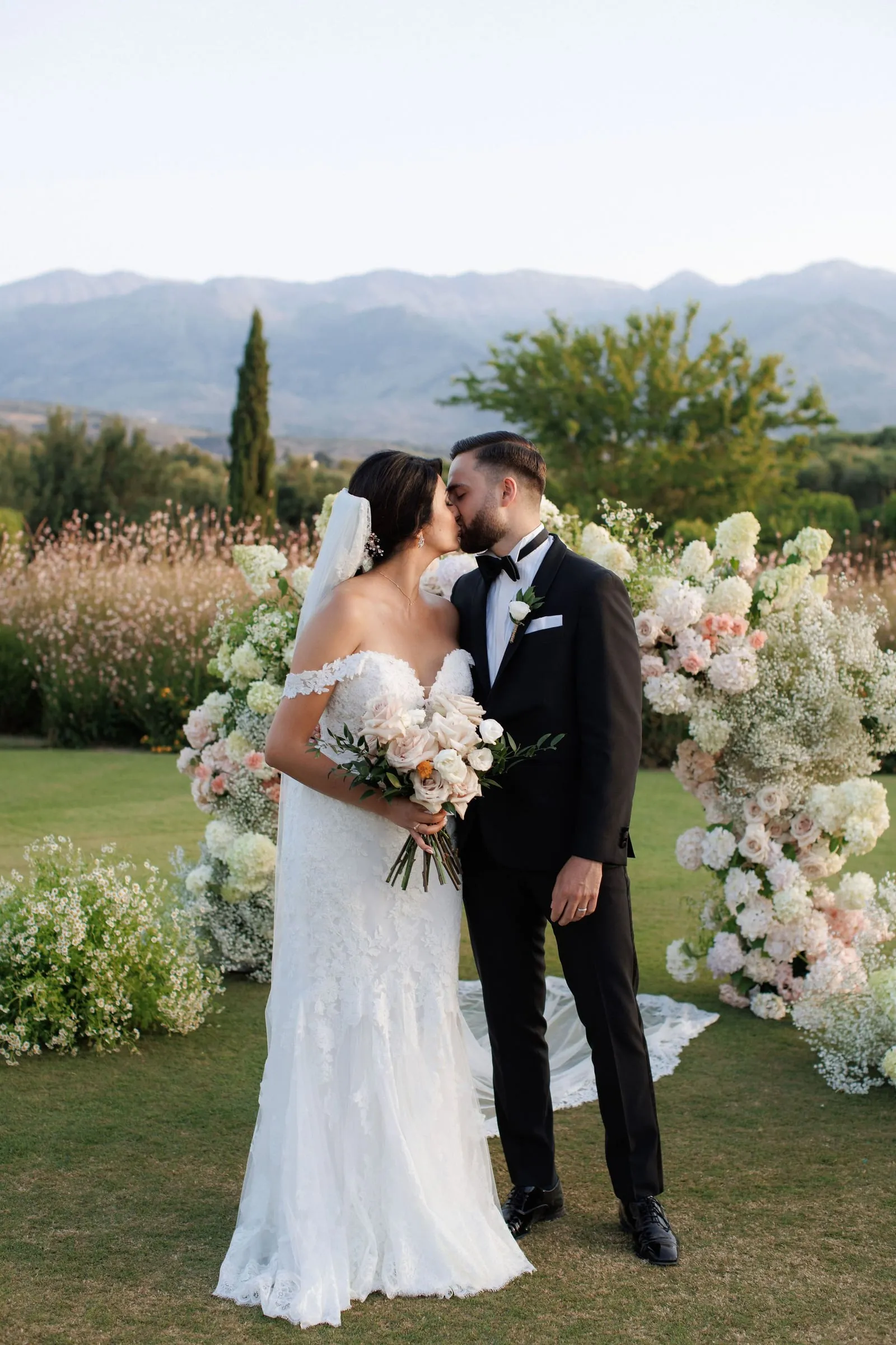 A bride in an off-the-shoulder lace gown and a groom in a black tuxedo kiss beneath a floral arch of cream and blush hydrangeas, roses and baby's breath on a lawn at Ktima Roxani in Crete, Greece. She holds a bridal bouquet of soft peach and cream roses with olive foliage, and a misty mountain range rises behind them under a pale evening sky. This is a wedding ceremony photo, with the flowers, arch and grasses framing the couple in a calm, romantic light.