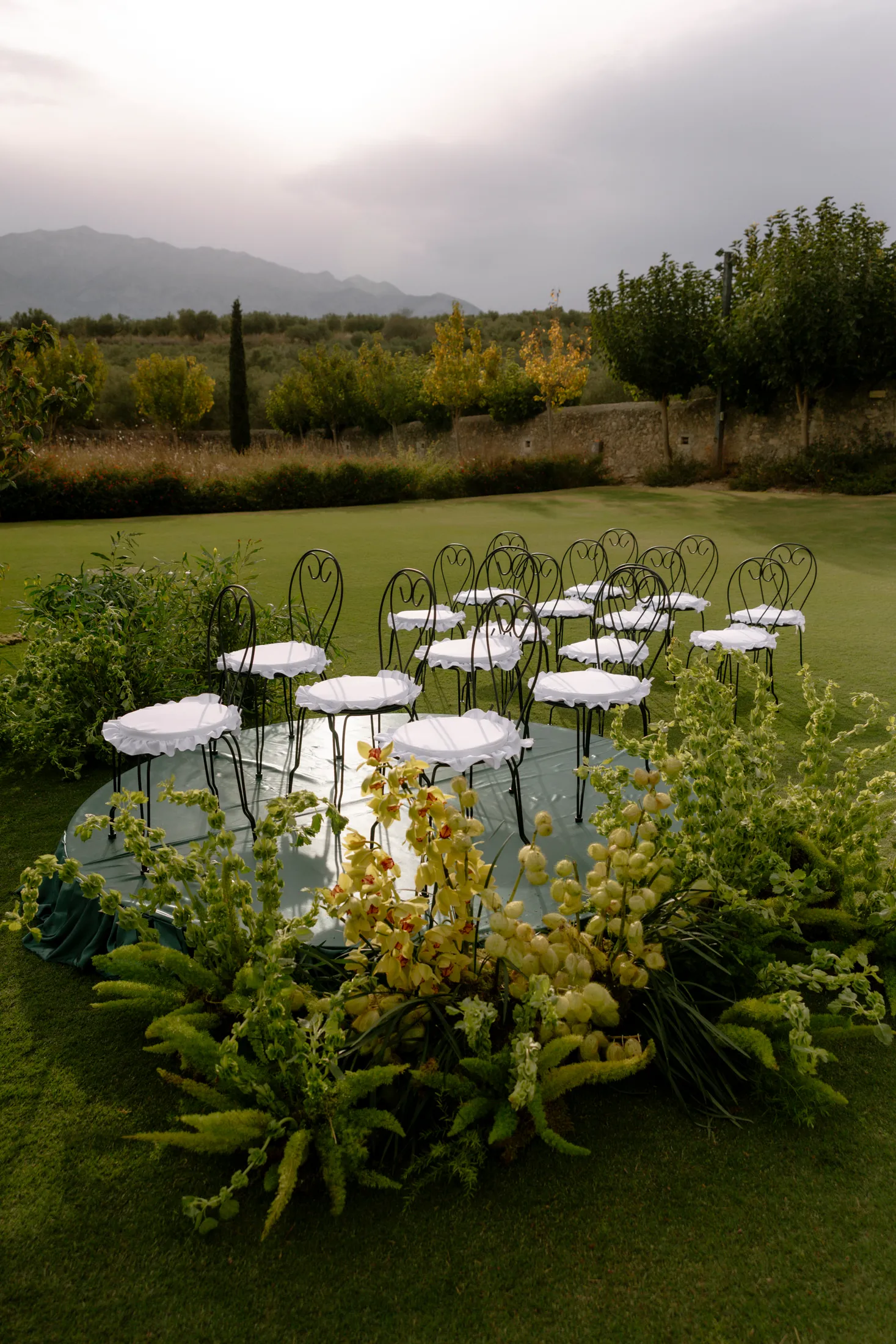 A circle of black wrought-iron chairs with white ruffled seat covers sits on a round green platform on a lawn, framed by lush greenery and tall pale yellow orchid spikes, with ferns and trailing foliage at the base. Behind, a stone wall, olive and round-headed trees, and misty mountains under a soft grey sky create a calm, green mood. This is a wedding ceremony setting at Ktima Roxani Estate in Crete, Greece.