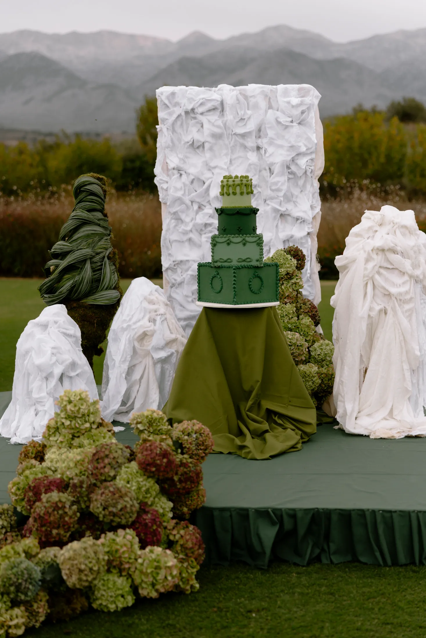 A tiered wedding cake in shades of sage and forest green sits on a draped green pedestal on a raised green platform, set on a lawn at Ktima Roxani in Crete, Greece. Clusters of green and burgundy hydrangeas spill across the foreground, and sculptural white fabric forms rise on either side, with a tall white draped panel behind and misty mountains beyond.