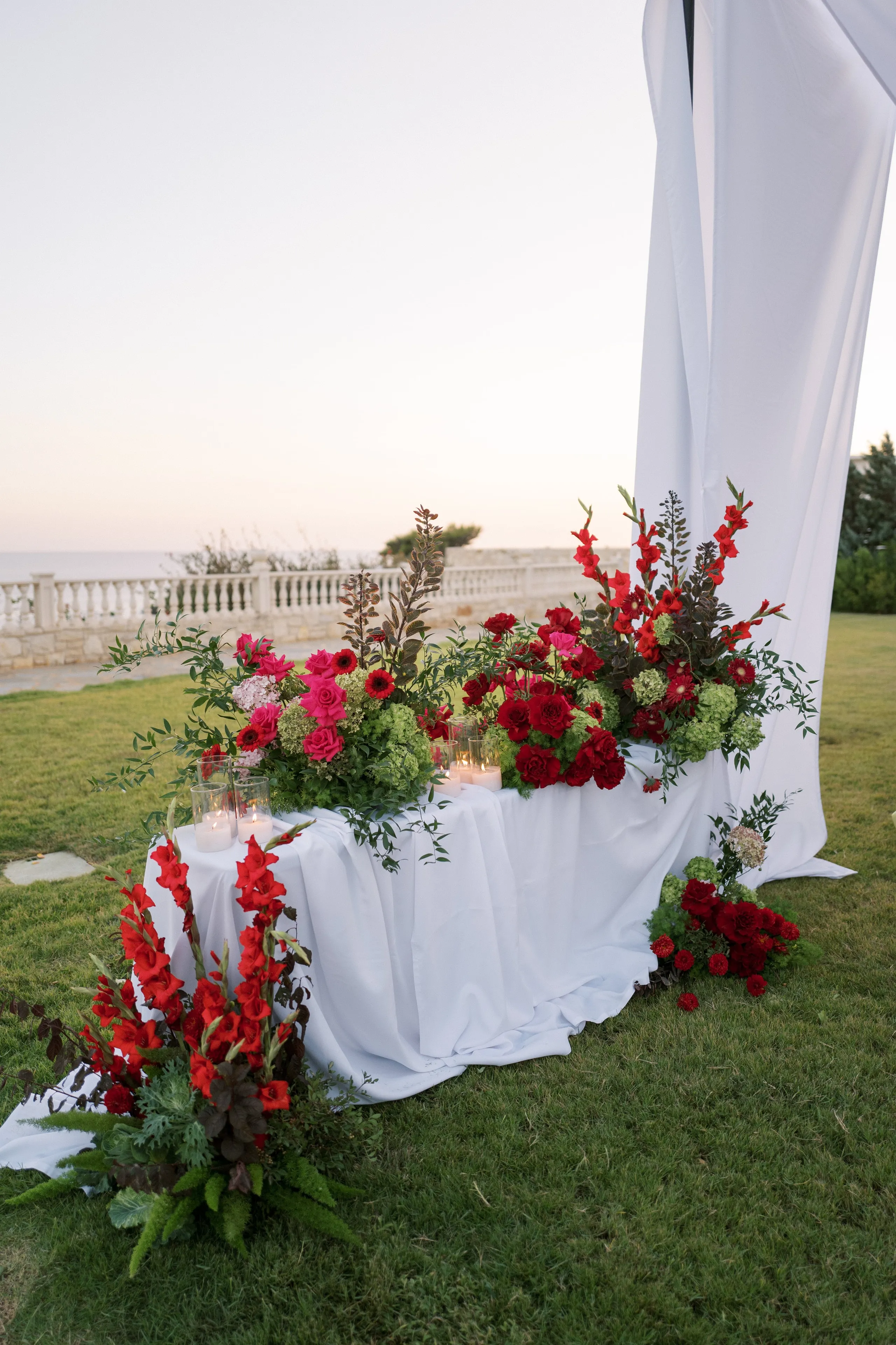 Wedding ceremony flowers in Crete, Greece, arranged on a long white draped table on the lawn at Wine Dark Sea Villas in Rodia. Two lush clusters of deep red roses, hot pink blooms, red gladioli and pale green hydrangeas spill over the cloth, with trailing greenery and dark bronze foliage, while tall glass hurricane candles glow at the centre. A white fabric panel rises behind, and a stone balustrade and the evening sea fade softly into a pale sky.