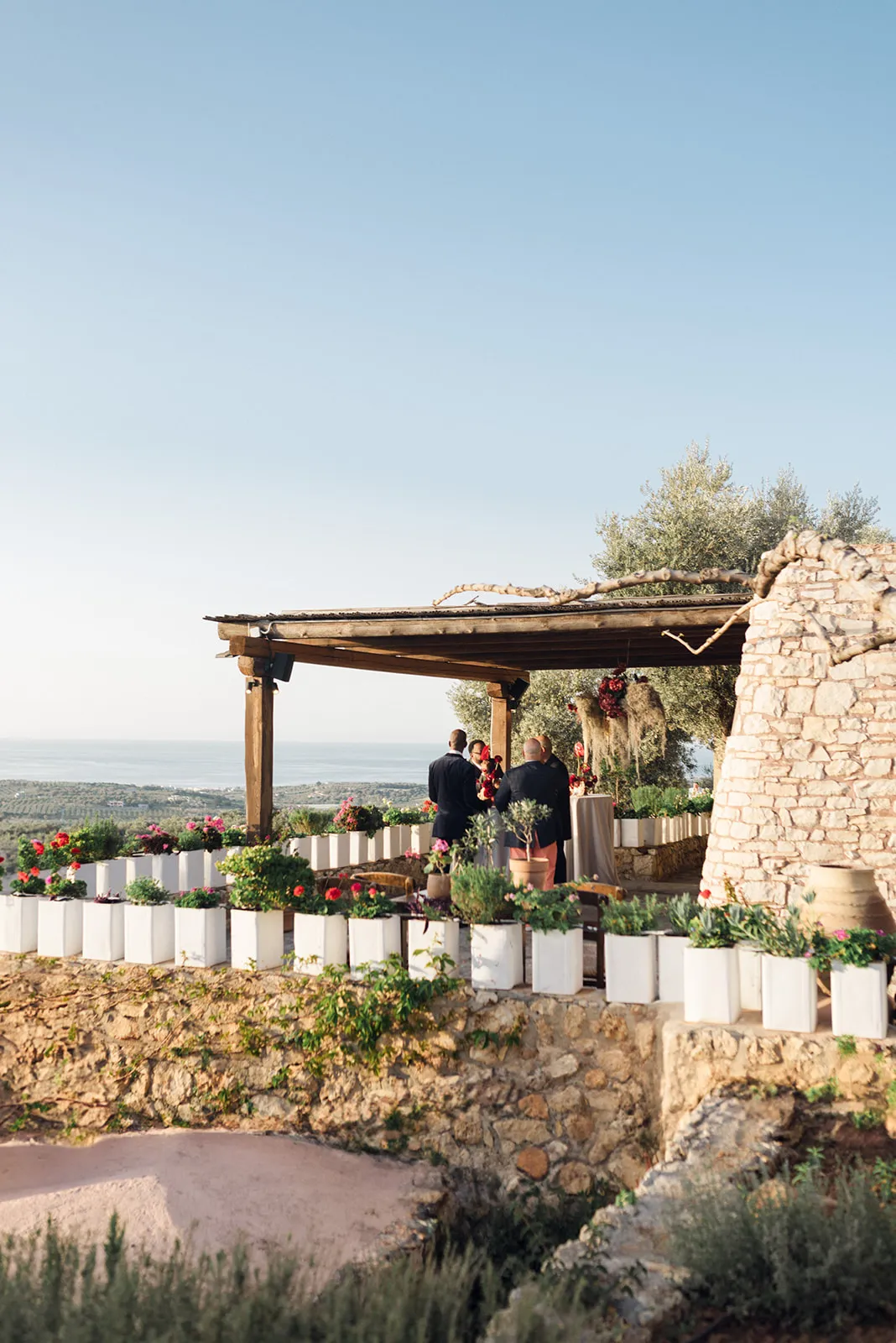 A wooden pergola on a hillside terrace at Agreco Farm in Greece, where two men in dark suits stand beneath a hanging arrangement of deep red blooms trailing dried moss. Along the stone wall, rows of white square planters overflow with pink and red geraniums and green shrubs, with the blue Mediterranean and olive groves stretching out below in the soft evening light.