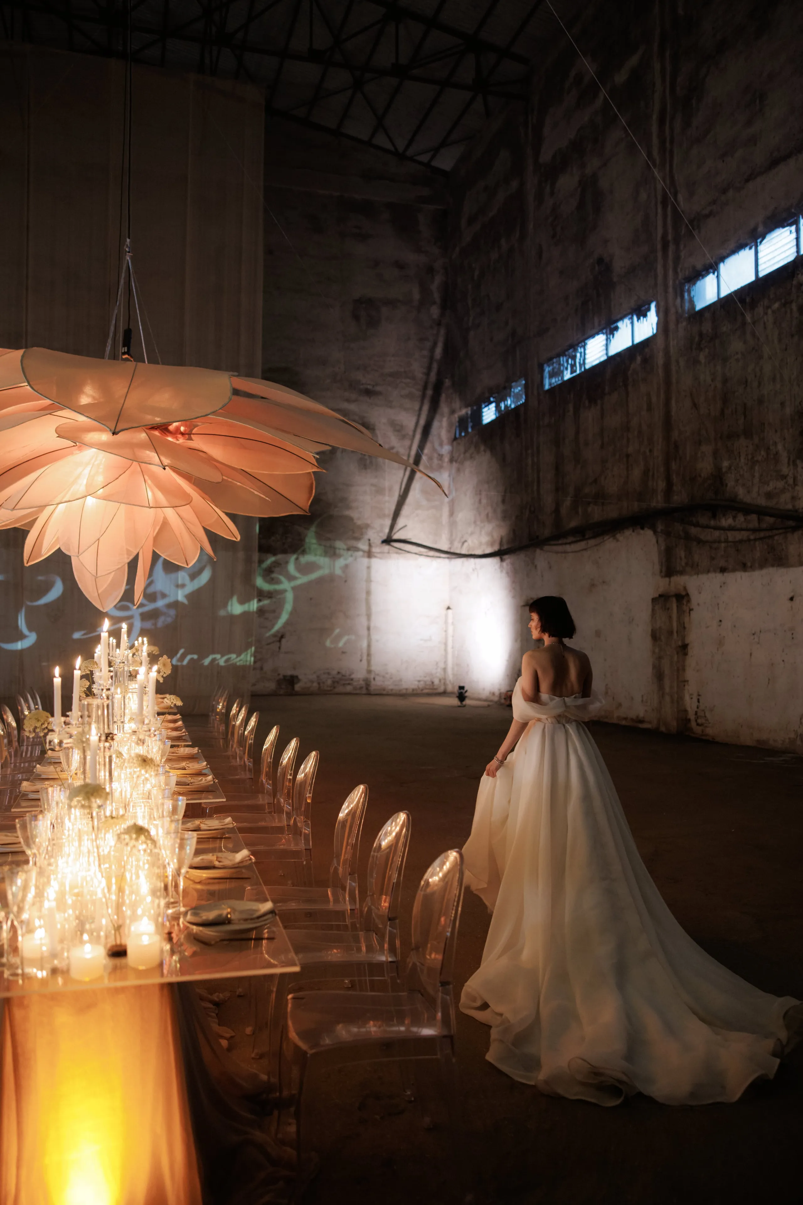 A bride in a softly structured ivory gown with a long, flowing train stands with her back to the camera in a vast, shadowy industrial hall, gazing down a long table lit by clusters of tall white candles and clear glass vases of pale blooms. Above the table hangs a large pink-peach paper lantern shaped like an open flower, and clear ghost chairs line the table. This wedding table styling is set in Rethimno, Crete, Greece.