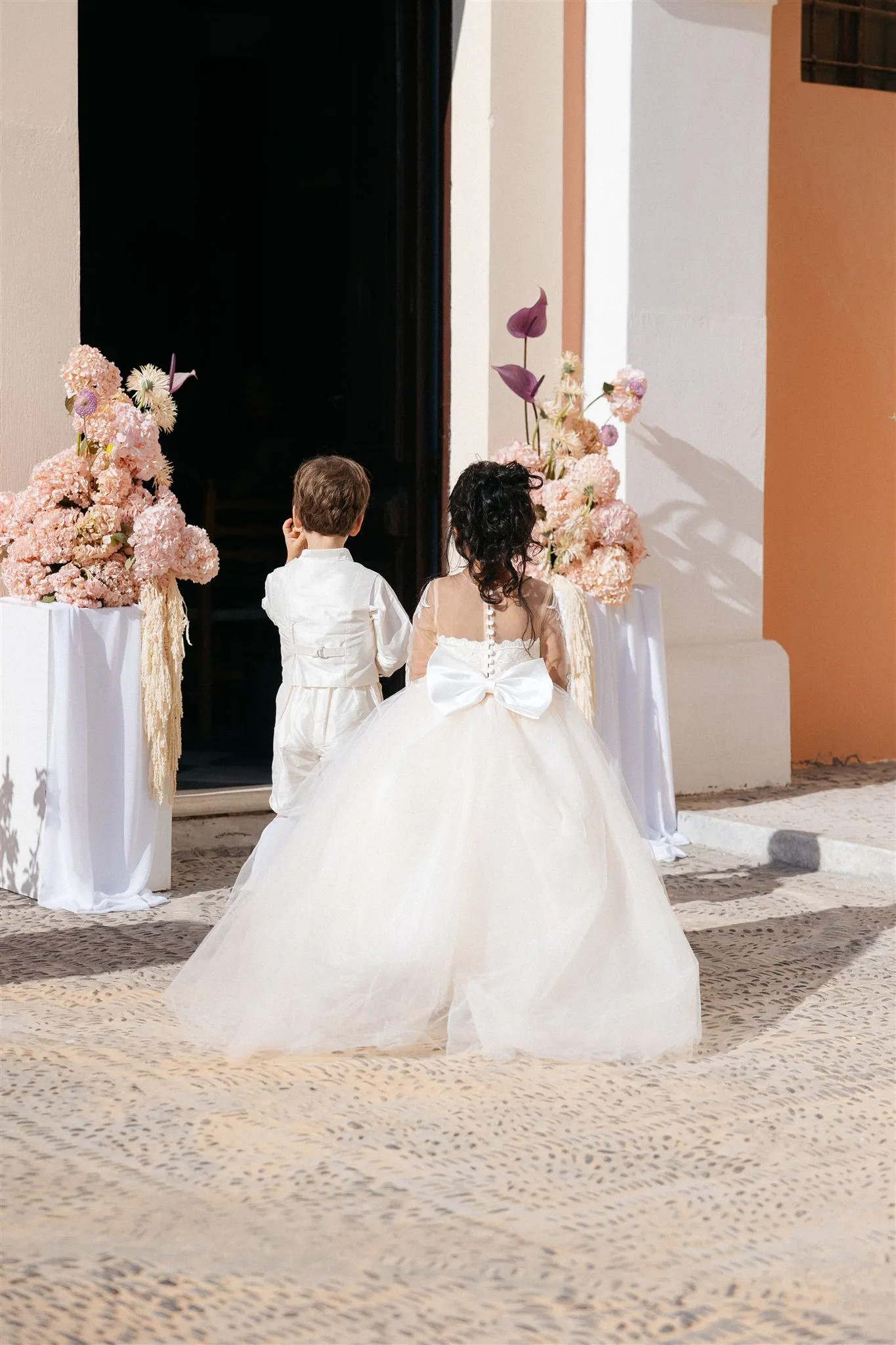Two young children in white stand at a dark doorway with their backs to us, a small boy in a cream suit and a girl in a full tulle gown with a satin bow at the back. Flanking the entrance are two tall floral arrangements on white draped pedestals, packed with blush-pink hydrangeas, pale cream blooms, and deep plum anthurium spikes. This wedding flower display is set at Islet Santorini in Thira, Greece, where warm sunlight falls across the patterned stone floor and the ochre wall.